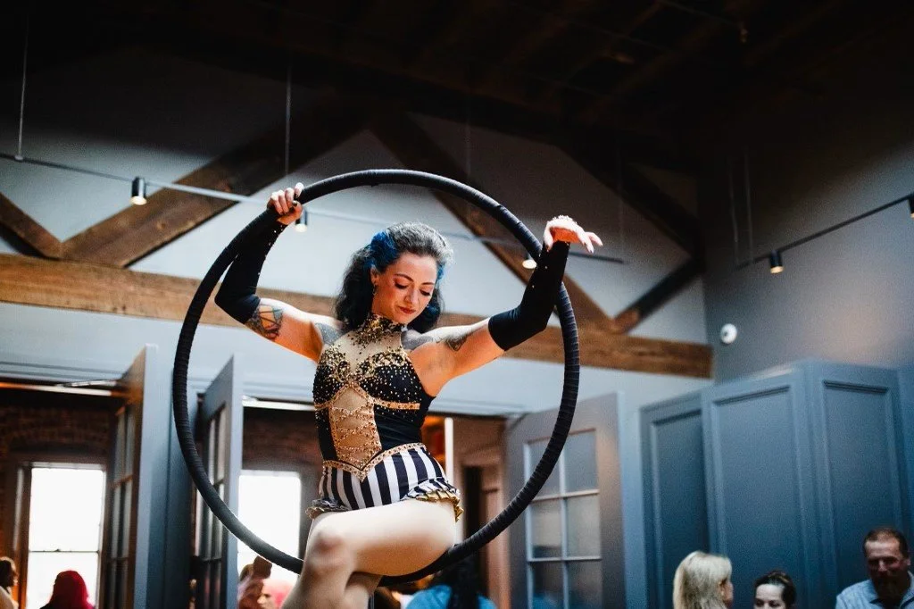 A woman performing a circus act with a hoop in an indoor venue with exposed wooden beams and blue walls.