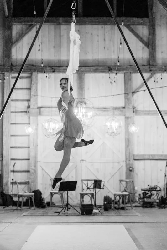 A female aerialist performing on sky ropes in a barn-like venue with wooden walls, chandeliers, and music stands.