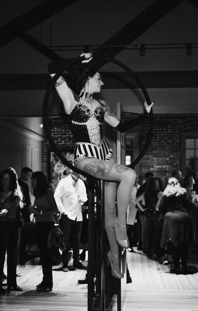 A woman performing aerial silk acrobatics inside a gym or studio, with people watching in the background.