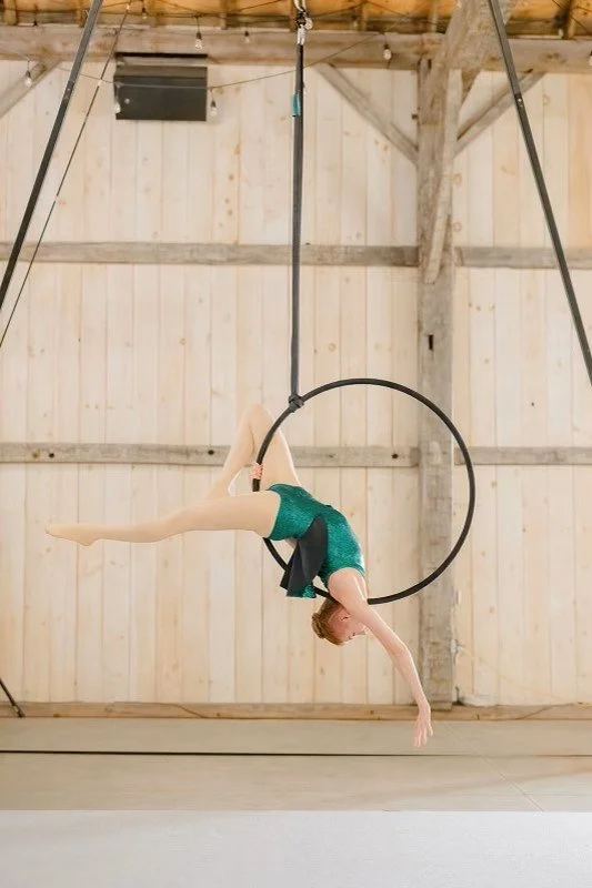 A gymnast performing on a hanging ring with one leg extended and the other bent, inside a wooden gymnasium.
