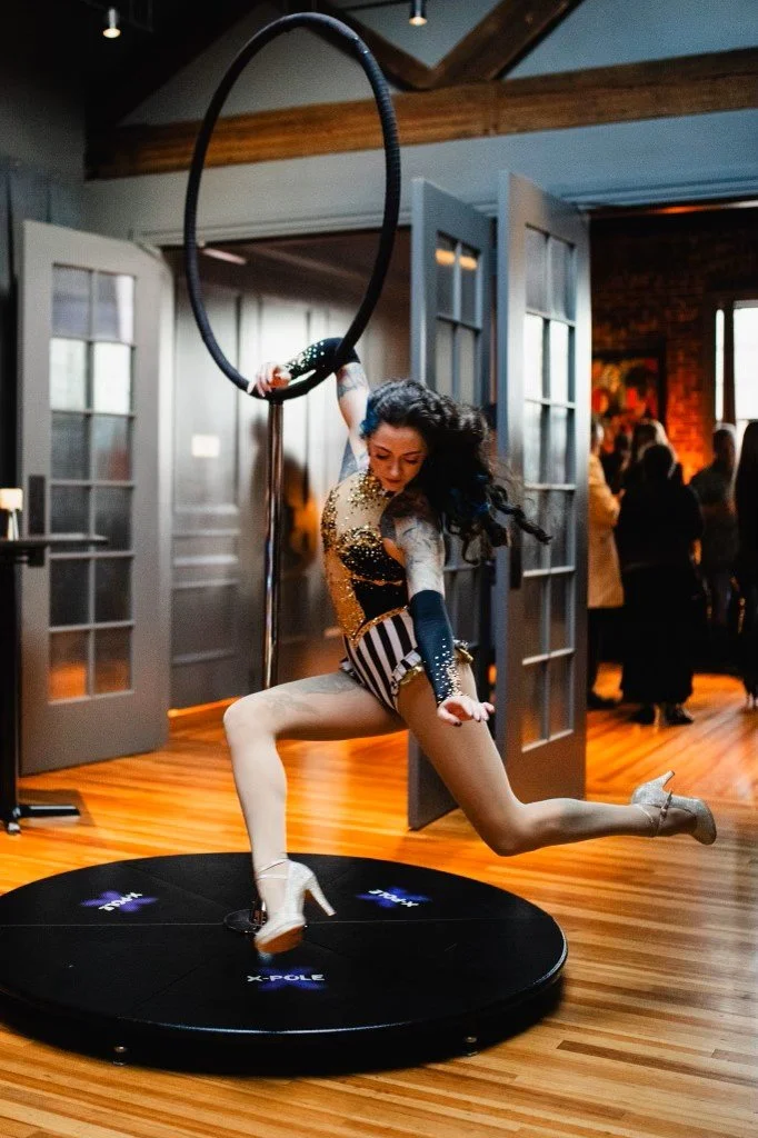 A woman performing an aerial dance on a hoop, jumping with one leg over a circular platform in a room with wooden floors and open doors, with a crowd in the background.
