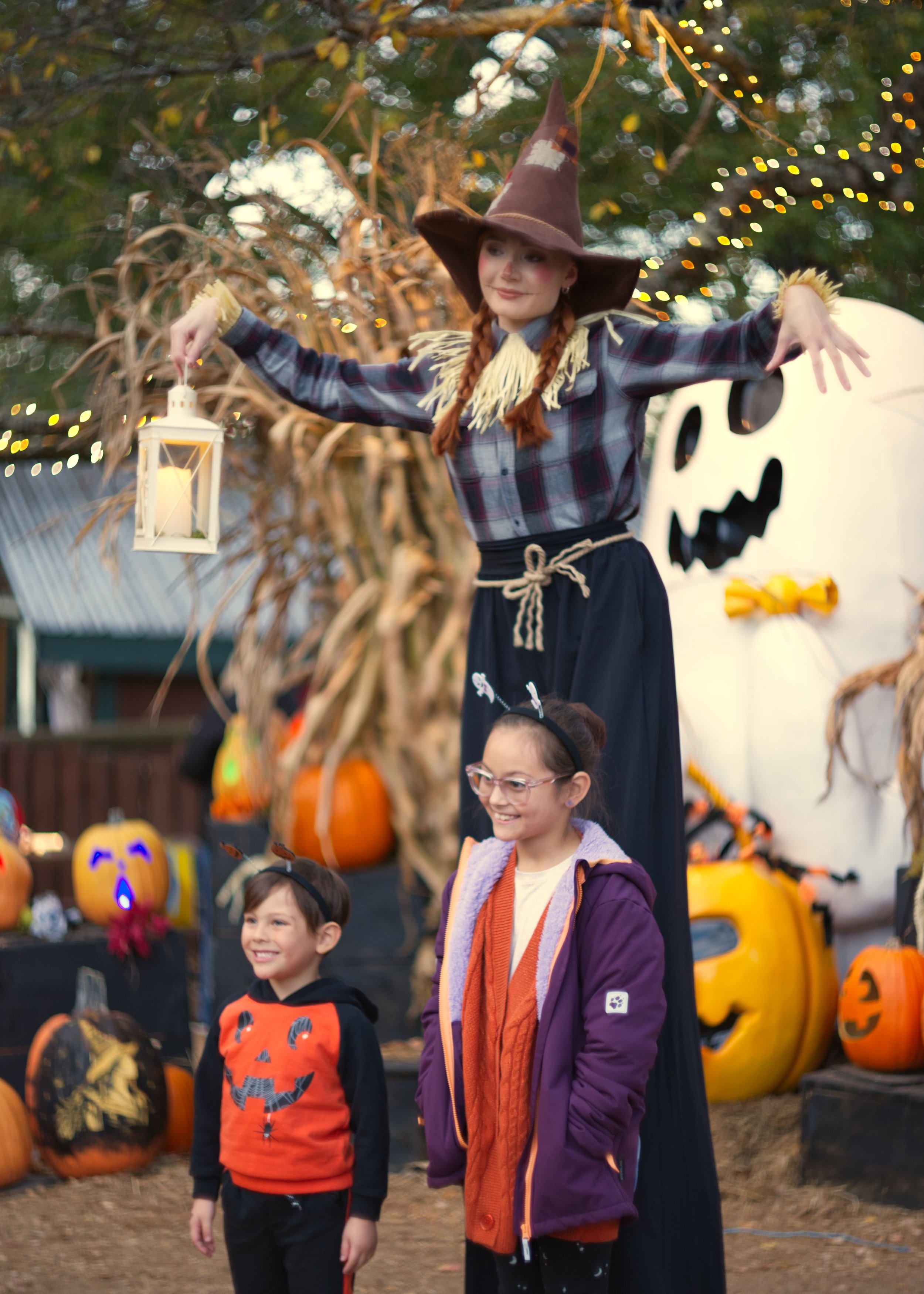 Three children at a Halloween event, two boys and a girl, standing in front of a large pumpkin with glowing faces. One girl is dressed as a witch with a purple coat, cat ears headband, and glasses. The other boy is wearing a pumpkin-themed sweatshirt