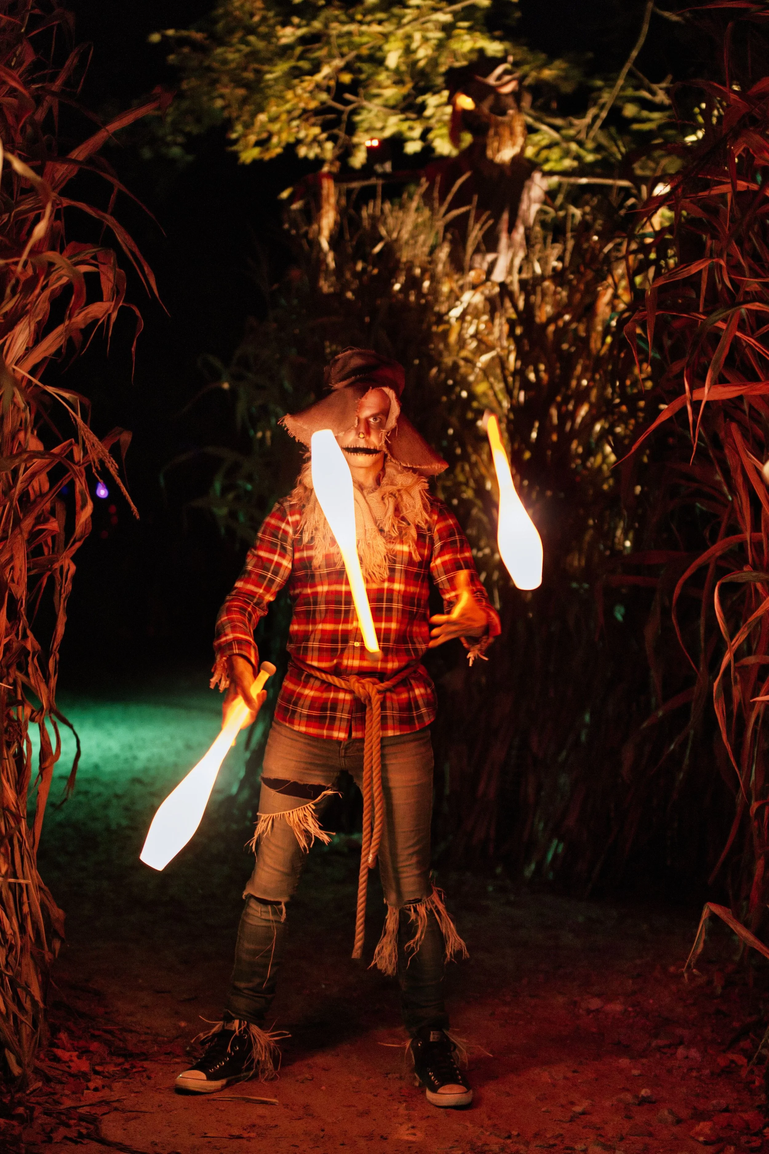 Person dressed as a scarecrow with a mask and plaid shirt, juggling flaming batons in a spooky, dark outdoor setting at night.