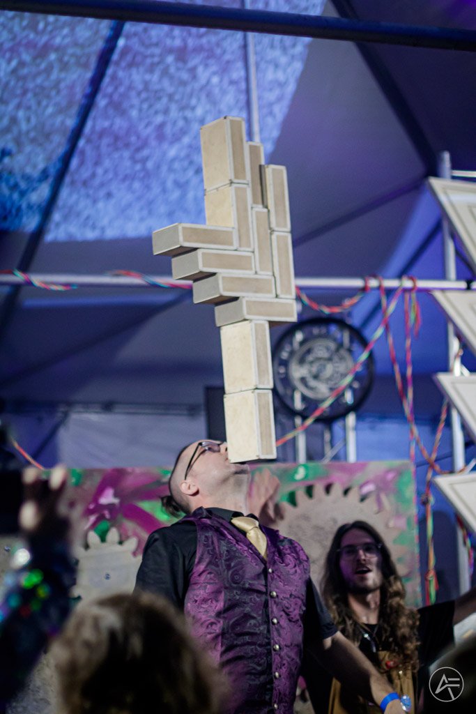 Person balancing a large Jenga tower on their face during a game or performance at an event, with spectators watching.