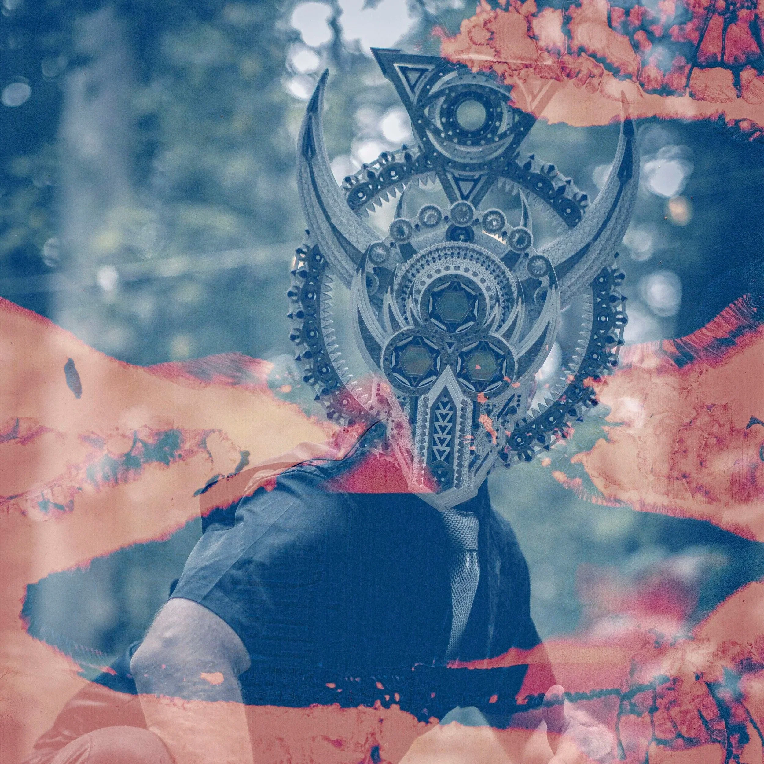Person wearing an intricate, metallic, geometric mask resembling a cosmic or mystical design, outdoors with a blurred background of trees and sky.