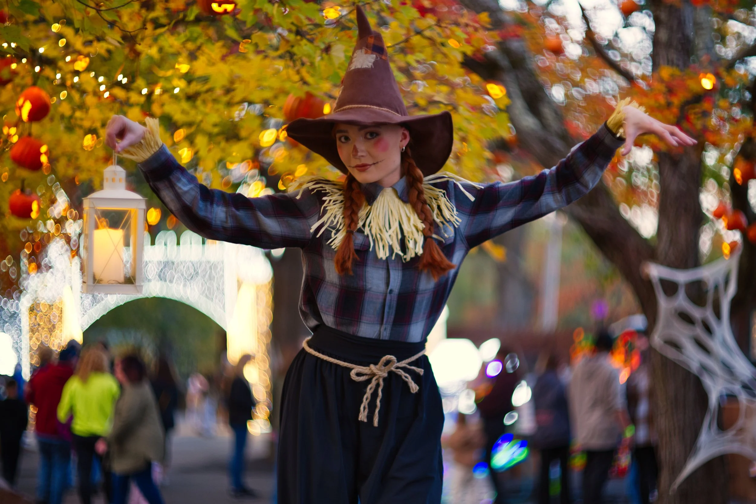 A woman dressed as a witch with a brown pointed hat, braided hair, and a plaid shirt with a straw-like collar, standing outdoors at night with Halloween decorations, such as a lantern, pumpkins, and cobwebs, in the background.