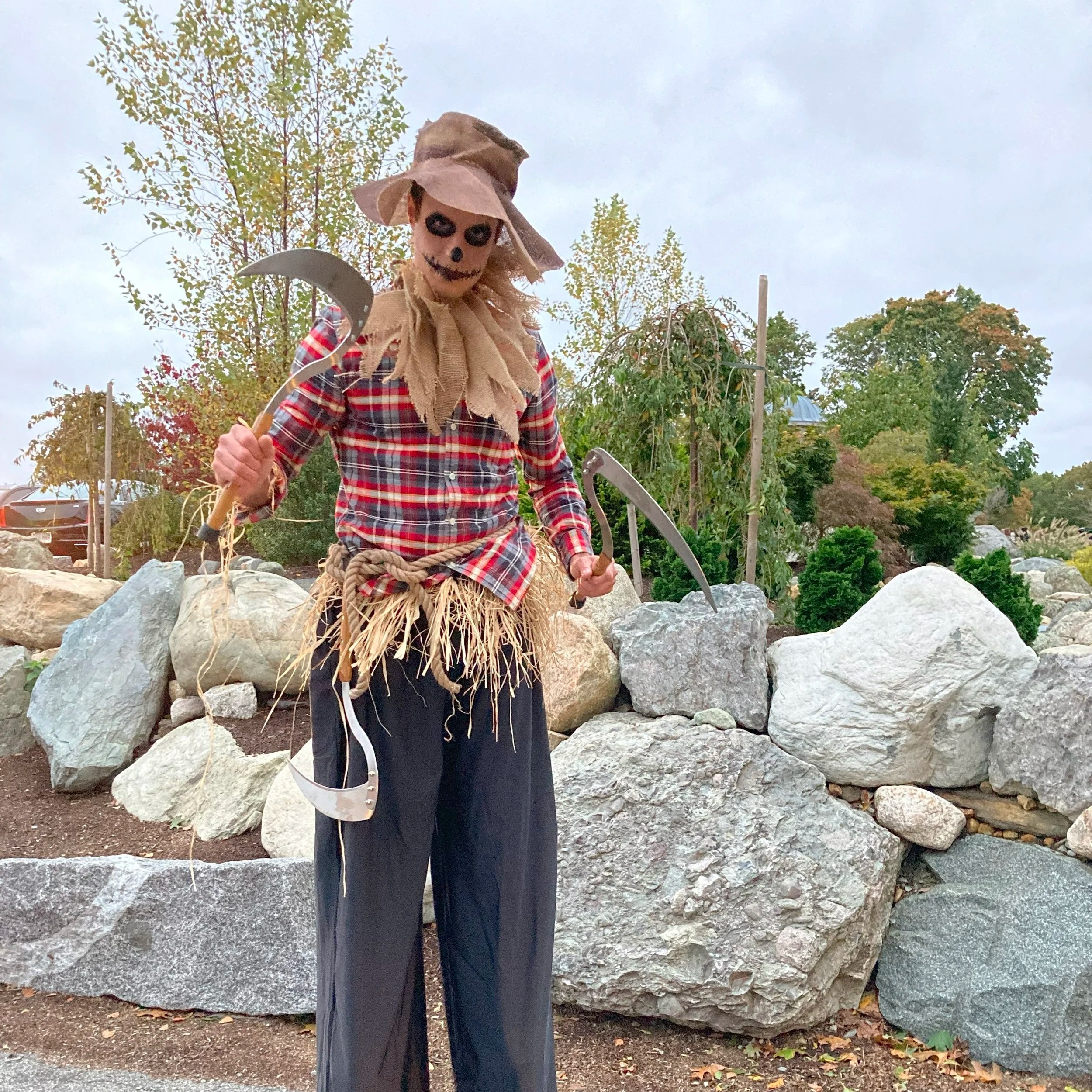 A person dressed in a scarecrow costume with a skull face painting, wearing a plaid shirt, straw skirt, and wide-brimmed hat, holding a scythe and axe, standing outdoors among rocks and trees.
