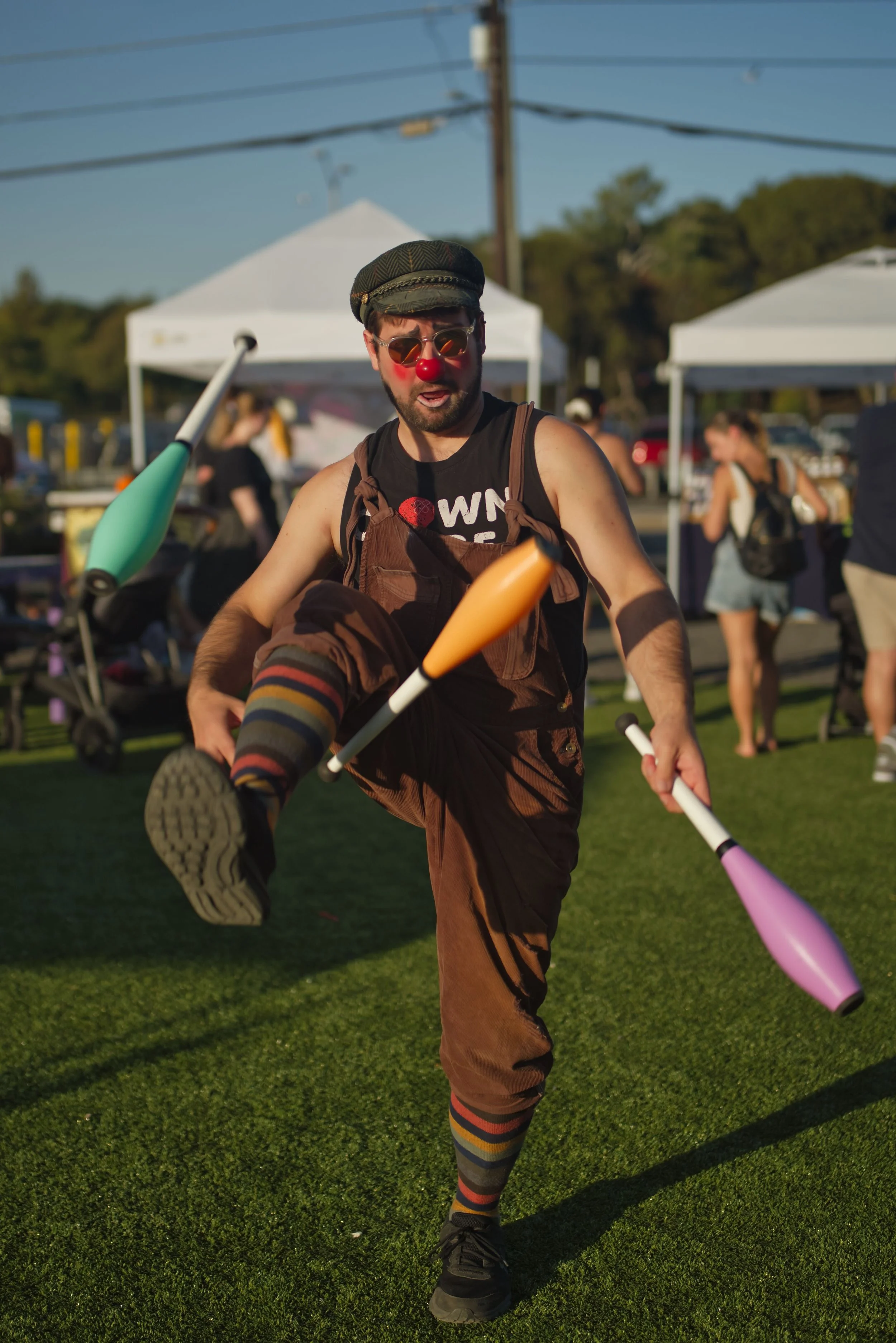 A man wearing a clown nose, sunglasses, and a cap juggles clubs at an outdoor event with tents and people in the background.