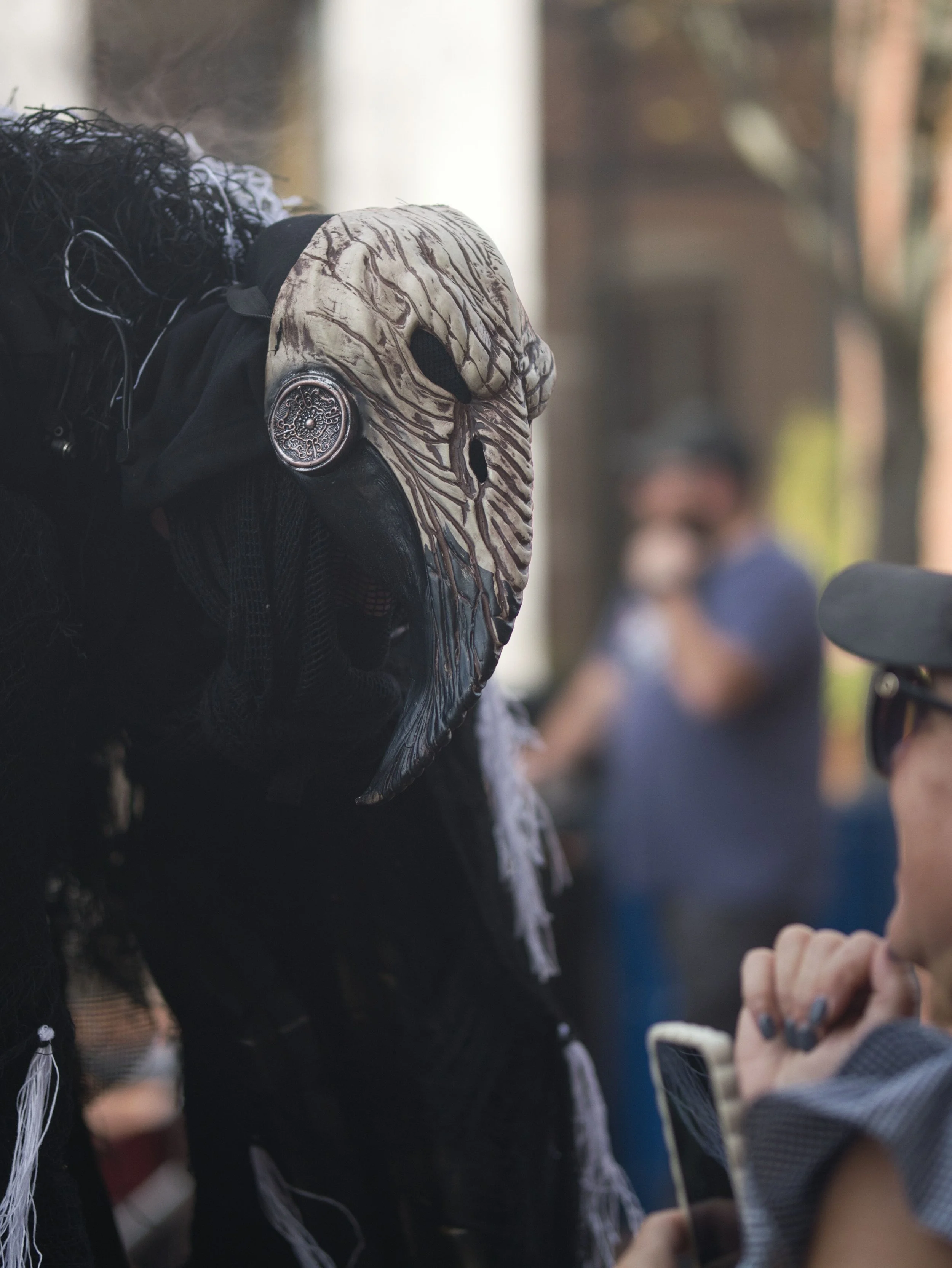 Person wearing a creepy, textured bird skull mask and dark costume, interacting with a woman taking a photo.