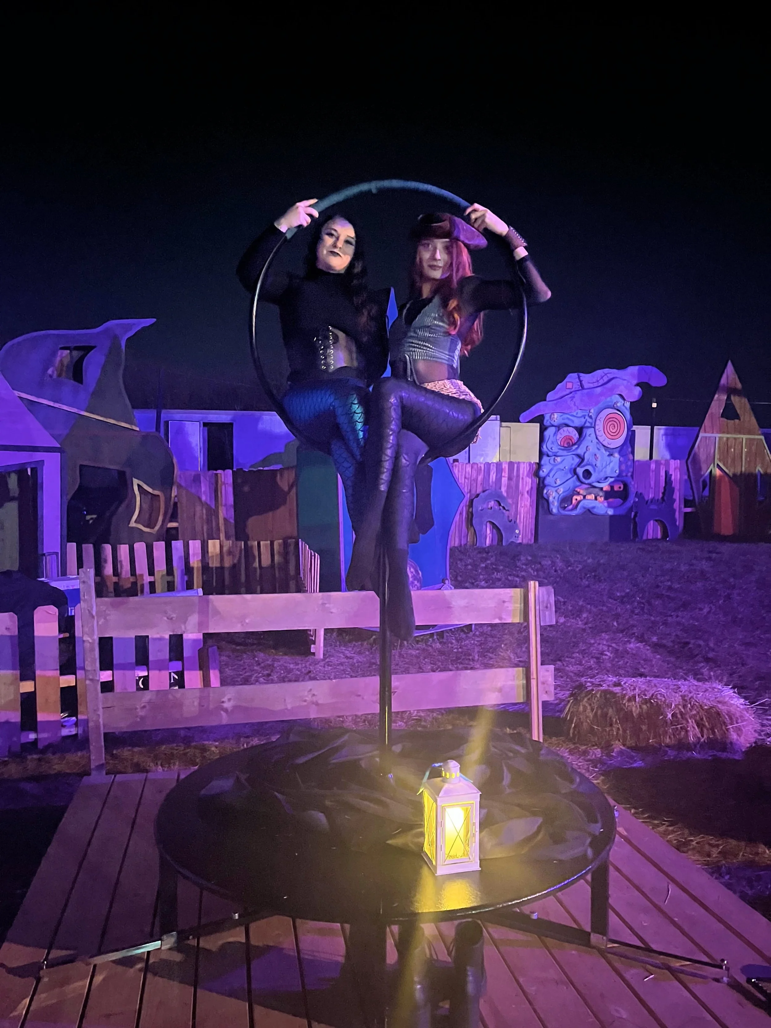 Two women dressed in dark clothing and fishnet stockings perform a balancing act on a pole in front of Halloween-themed decorations, illuminated by purple and yellow lights at night.