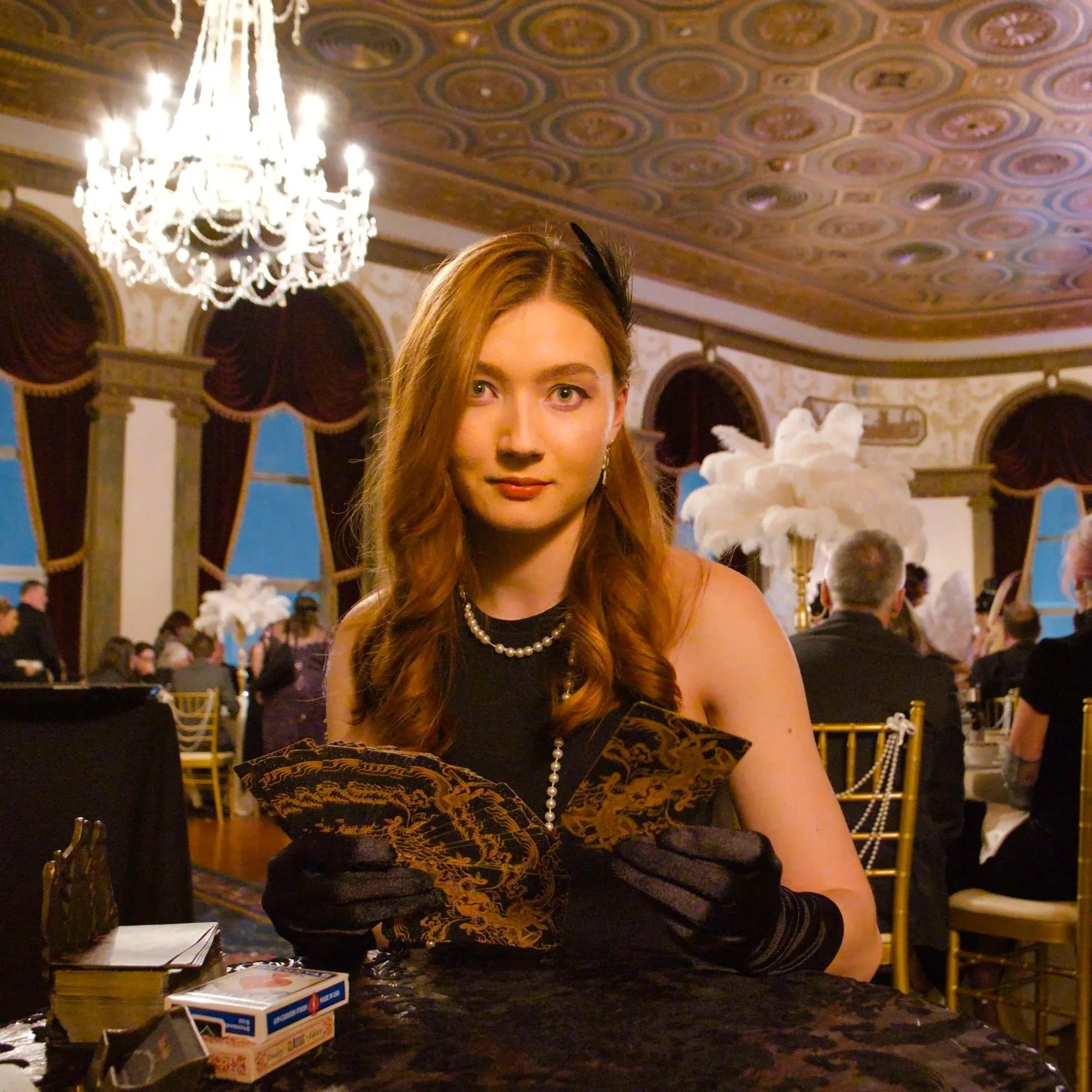A woman with long red hair, wearing black gloves, a pearl necklace, and black dress, sitting at a decorated event with chandeliers, large curtains, and tables with white feather centerpieces.