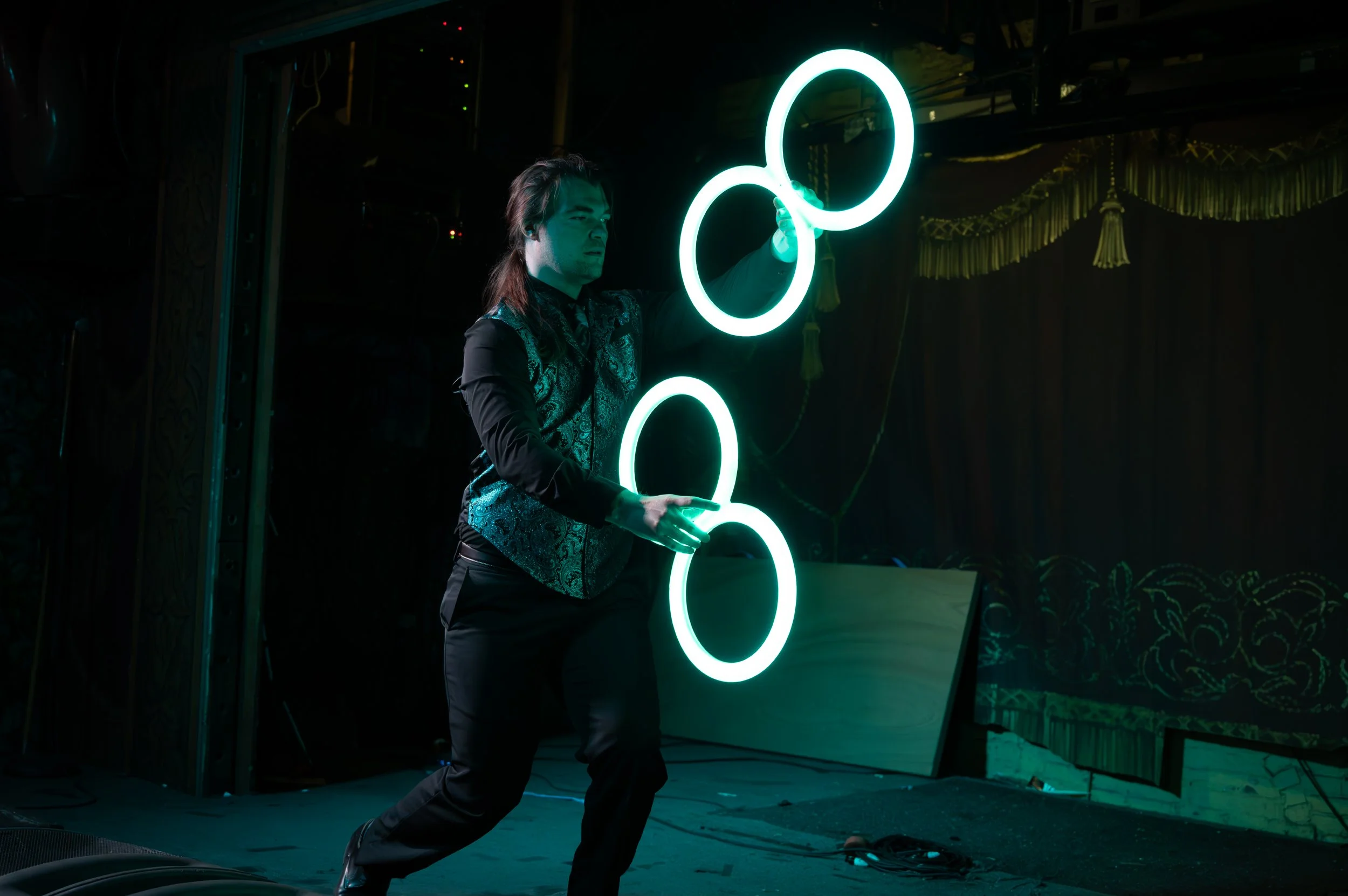 A performer on stage juggling neon-lit rings in a dark setting with stage curtains and equipment in the background.