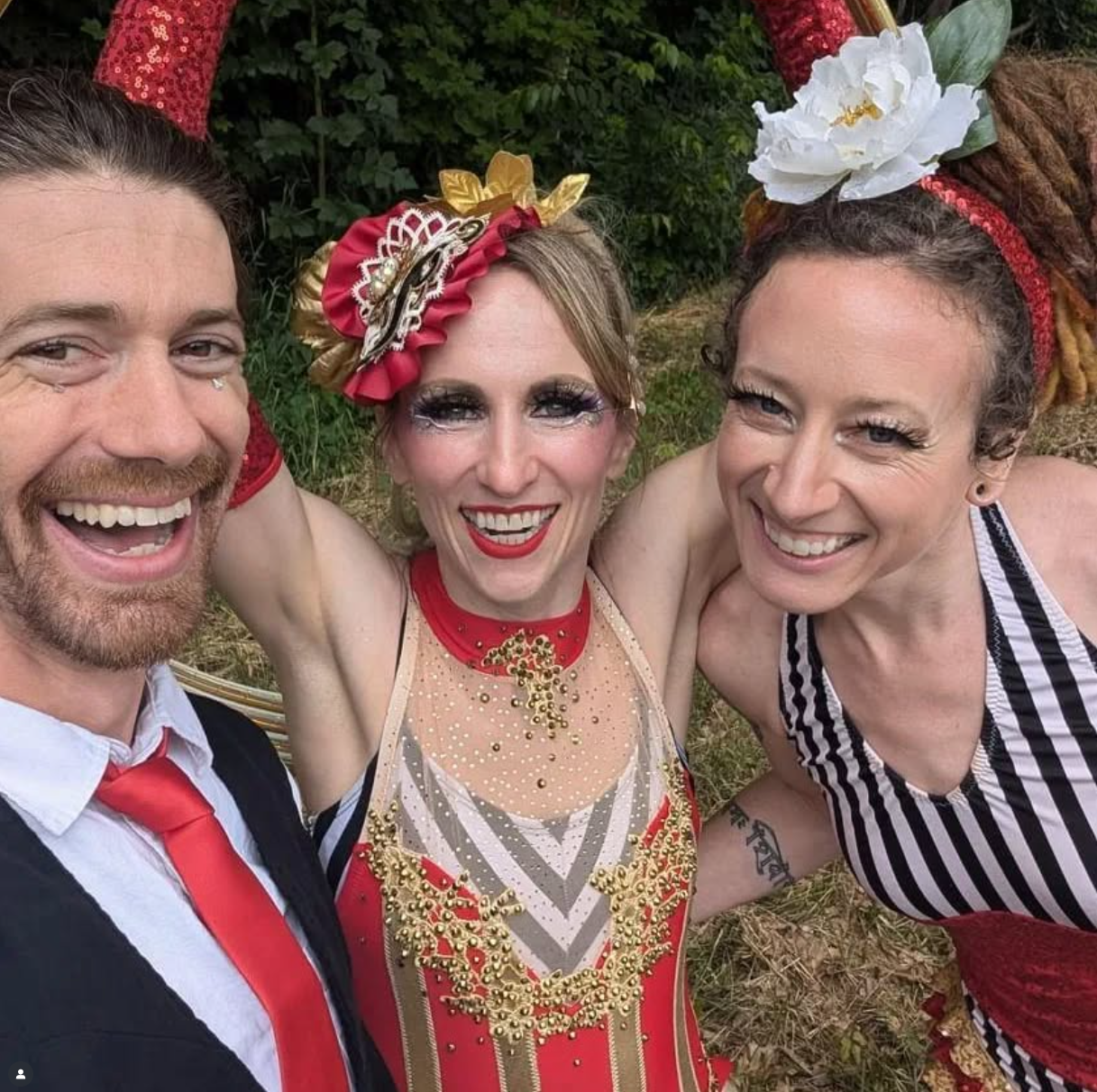 Three smiling people taking a selfie outdoors, dressed in colorful costumes with accessories and makeup, with greenery in the background.