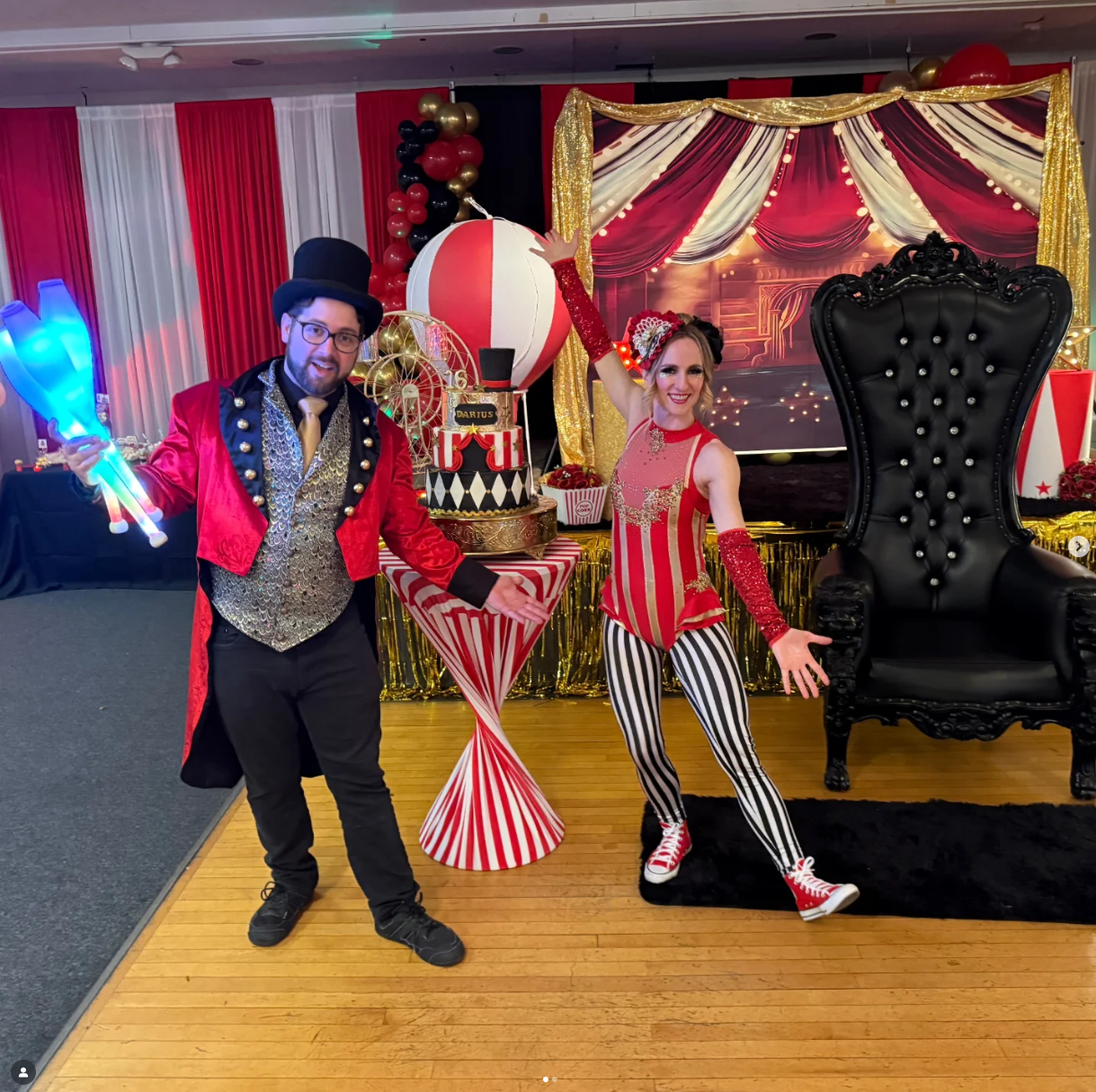 A man and woman dressed as circus performers standing in front of a circus-themed backdrop with a large black throne. The man is wearing a red jacket, silver vest, black top hat, and glasses, holding illuminated blue circus props. The woman is wearing a red and gold striped costume with striped black-and-white pants, red glittery gloves, and red sneakers, with her arms raised and smiling.