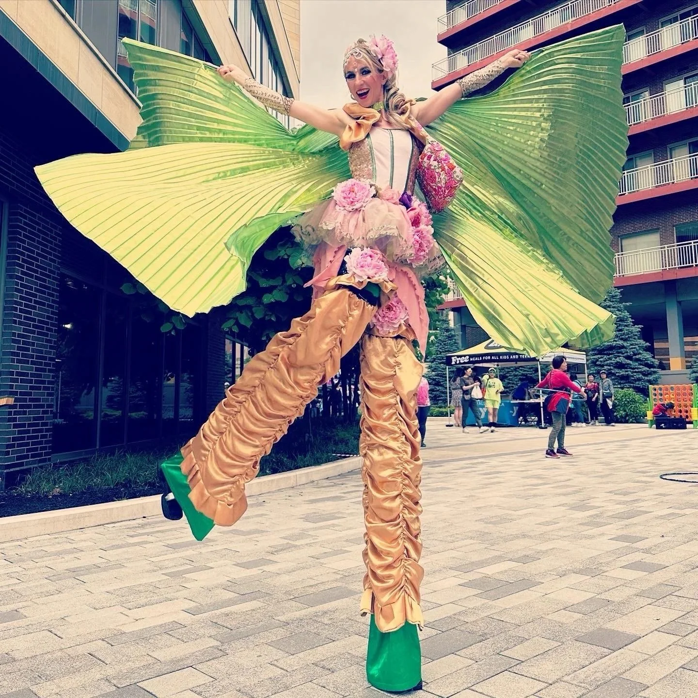 Performer on stilts dressed as a butterfly with green wings, wearing a pink and beige floral costume, in an outdoor urban area with people and buildings in the background.