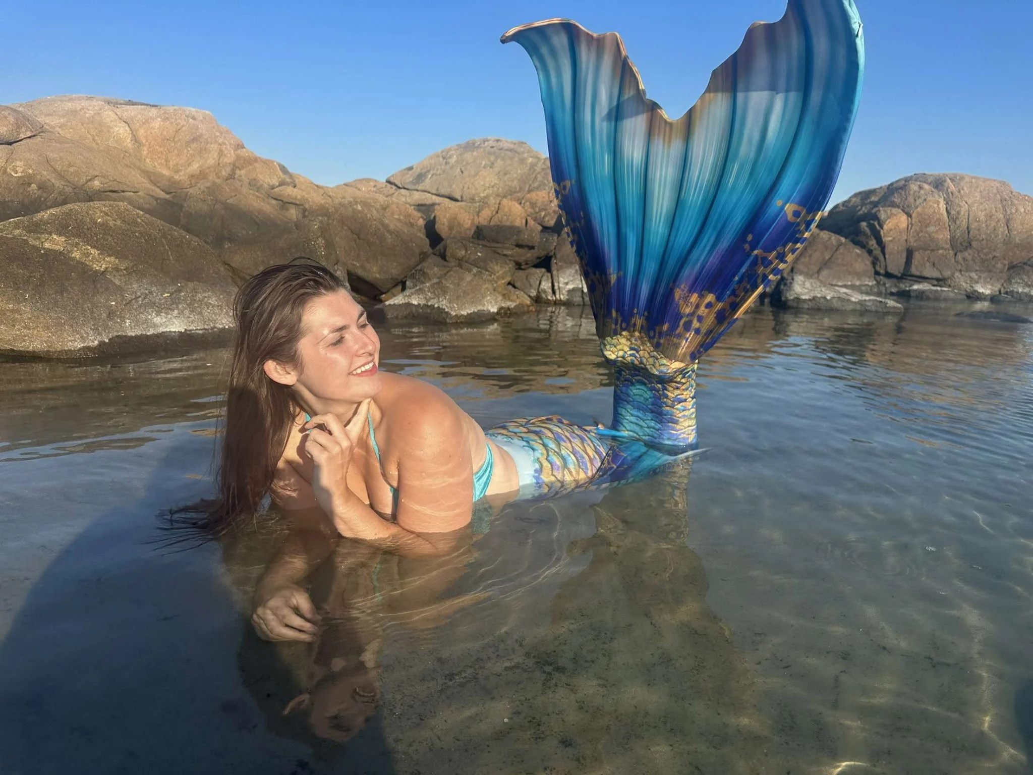 A woman in a mermaid costume with a blue and green tail is lying in clear water near rocks, smiling with her eyes closed, during daytime sunlight.