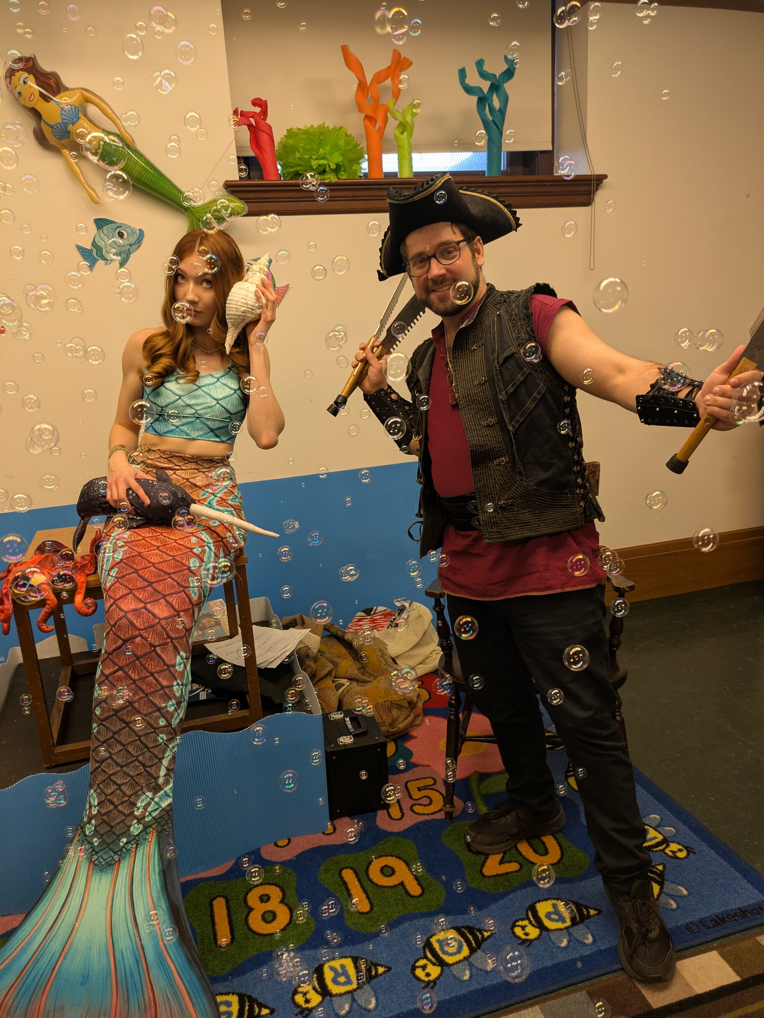 Two people dressed in mermaid and pirate costumes posing with bubbles. A woman with long red hair in a mermaid costume holds a seashell to her ear, and a man in pirate attire holds a toy axe. The background includes animated sea creature decorations 