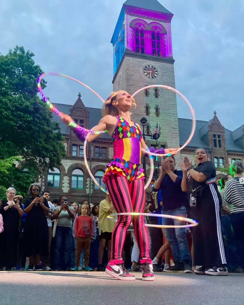 A female performer in colorful, patterned circus attire twirls glowing hoops on an outdoor street, with an audience and a historic clock tower in the background.
