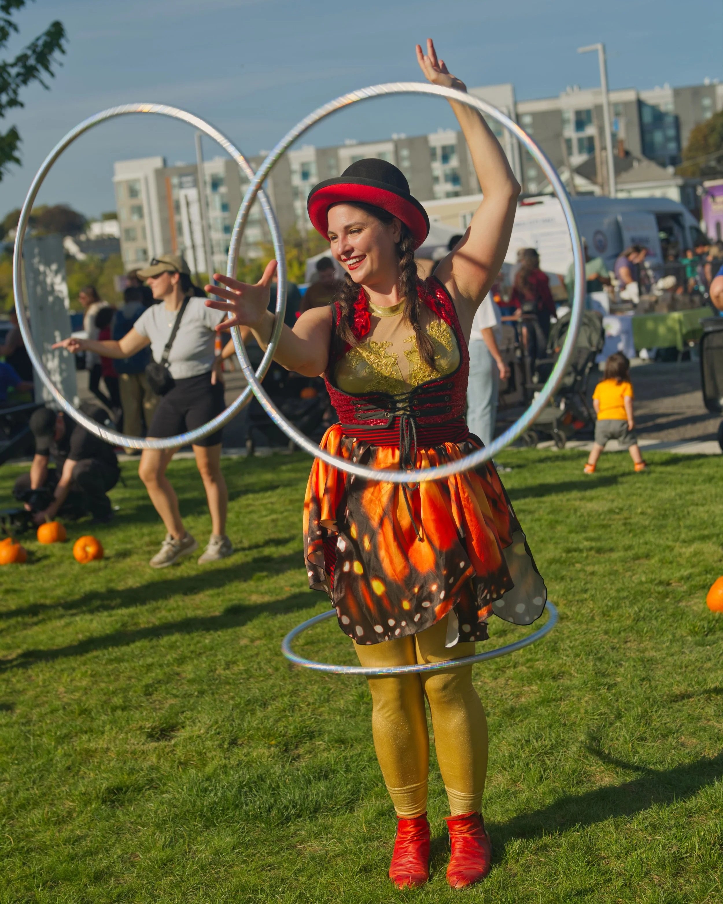 A woman smiling and performing with three hula hoops at an outdoor event during daytime, with other people and pumpkin decorations in the background.