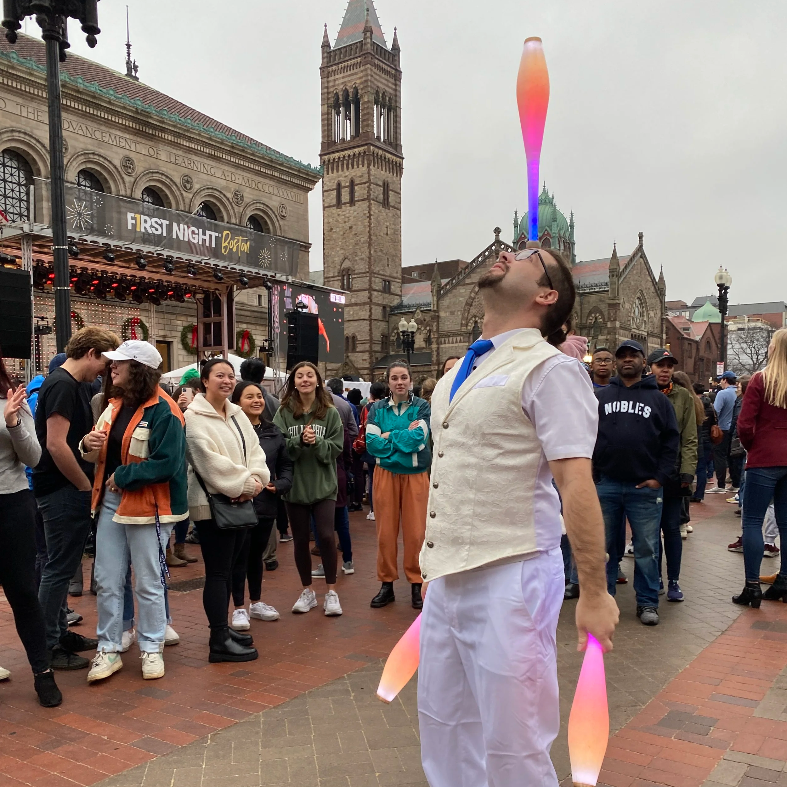 A street performer balancing a glowing object on his nose while spectators watch at an outdoor event, with a historic building and a church in the background.