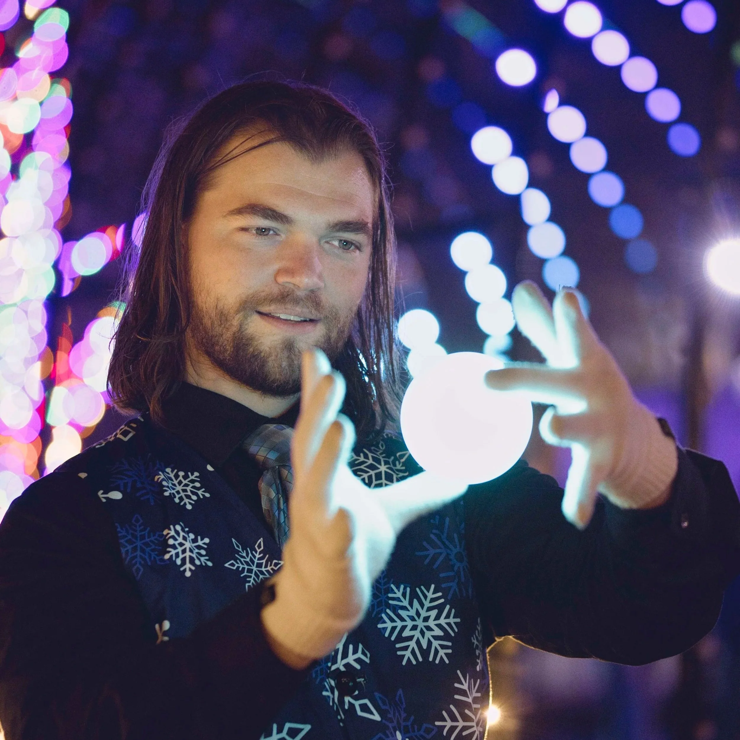 Man with long hair and beard holding illuminated orb at night with festive colorful lights in background