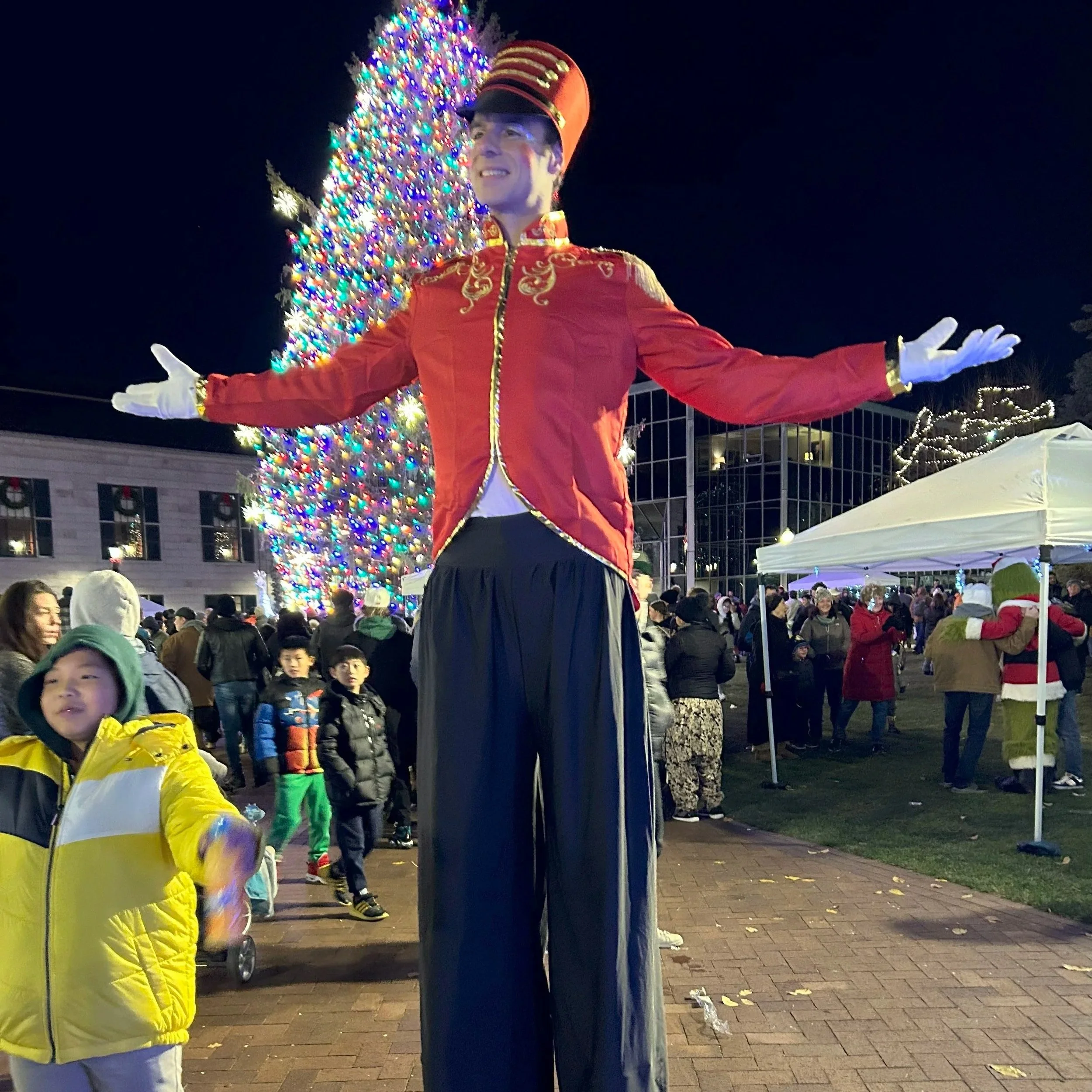 A person dressed as a circus or parade performer on stilts wearing a red jacket with gold embellishments, black pants, and a tall red hat, standing with arms outstretched in front of a large, decorated Christmas tree at night during a holiday celebration.
