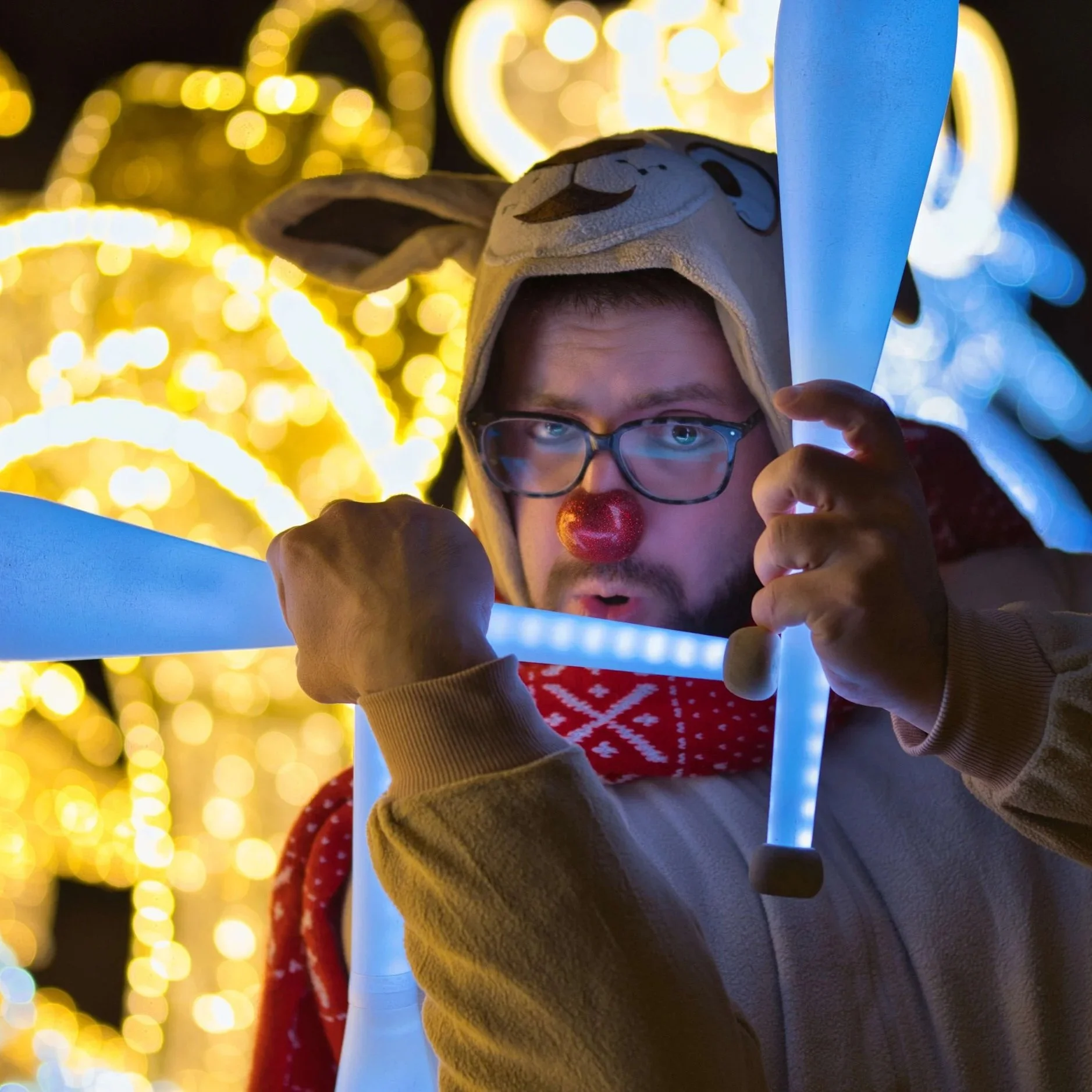 Person dressed in a reindeer or reindeer hat costume with glasses, a red nose, and a red patterned scarf, holding a glowing blue stick, with bright yellow and white holiday lights in the background.