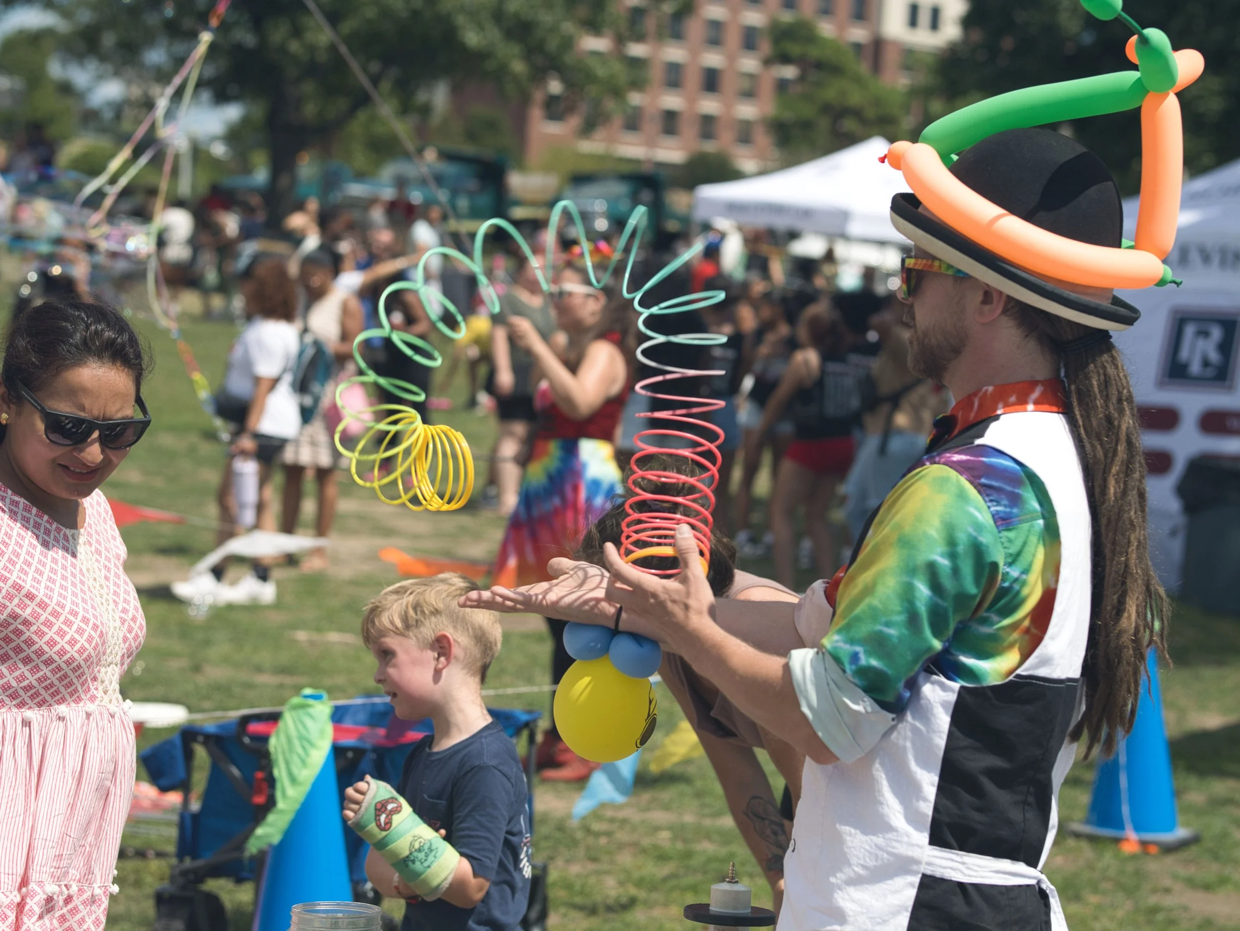 Man wearing a colorful tie-dye shirt, a hat with balloon animals and spirals, and sunglasses, performing a balloon twisting act at an outdoor festival with a crowd in the background.