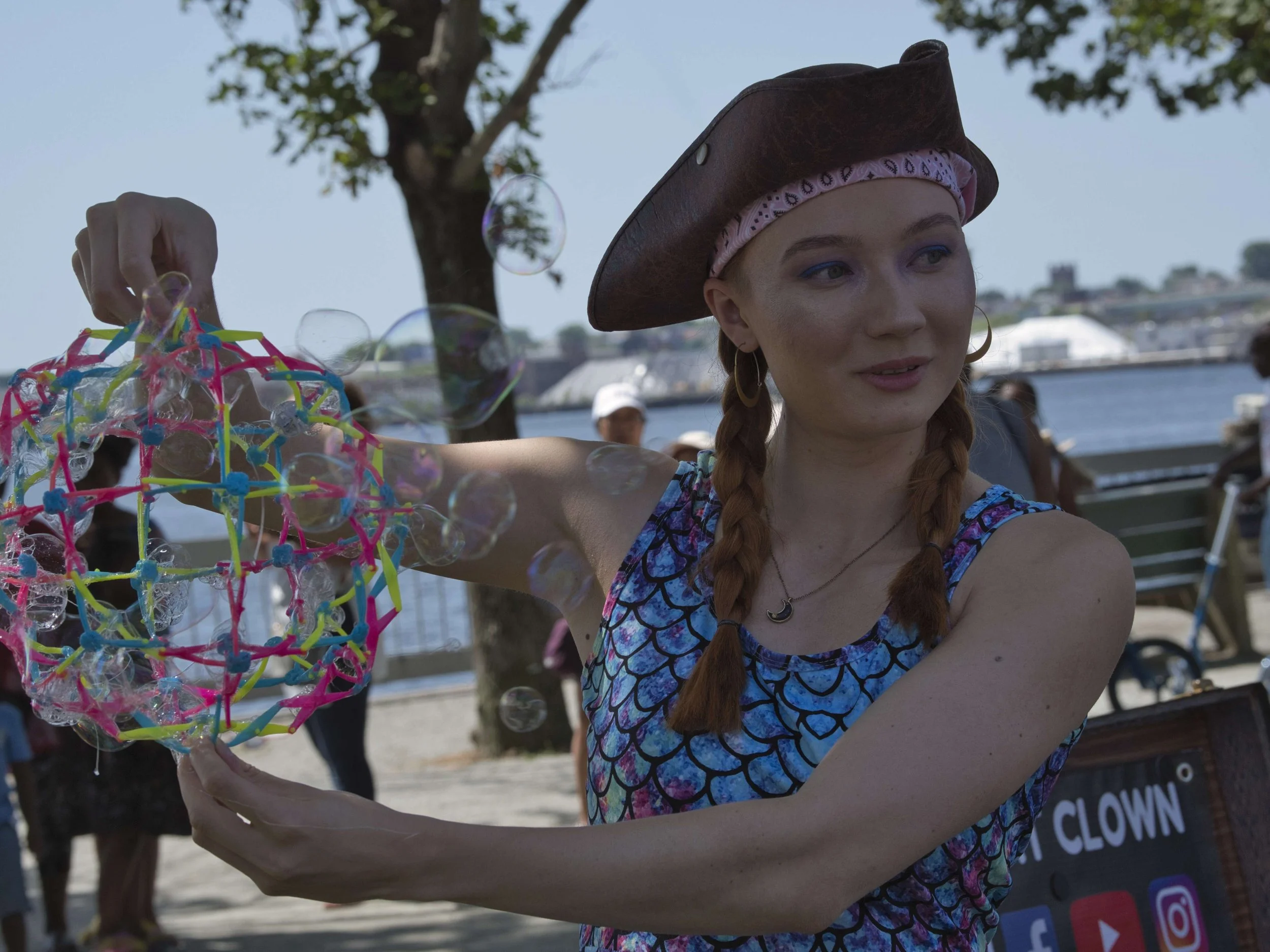 A young woman wearing a pirate hat and a blue patterned dress, is blowing bubbles outdoors near a body of water, with trees and other people in the background.