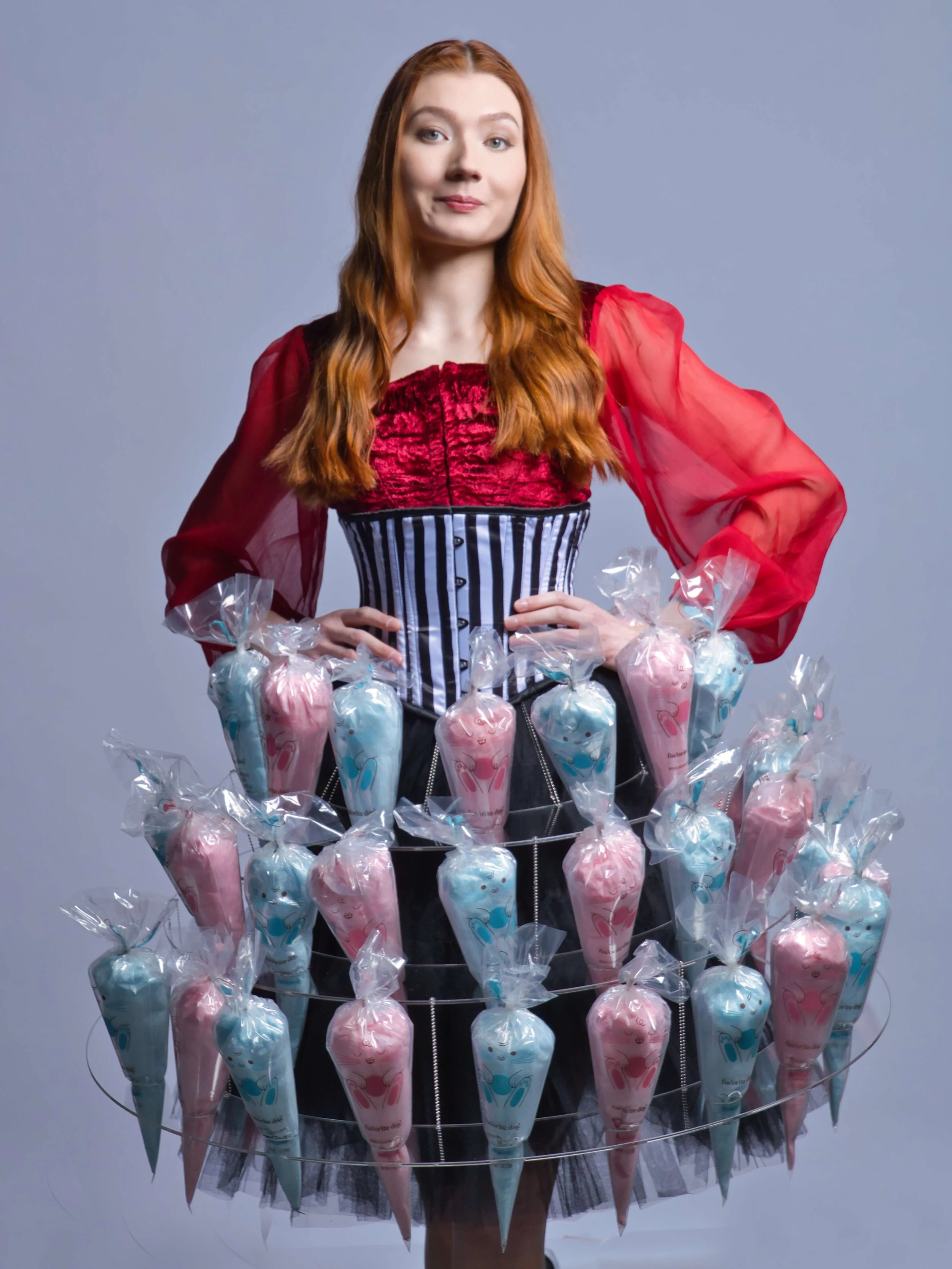 A woman with long red hair wearing a red and black corset with sheer red sleeves, standing with her hands on her hips, in front of a light gray background. She has a playful expression. In front of her, there is a circular candy display filled with pink and blue marshmallow candies wrapped in clear plastic.