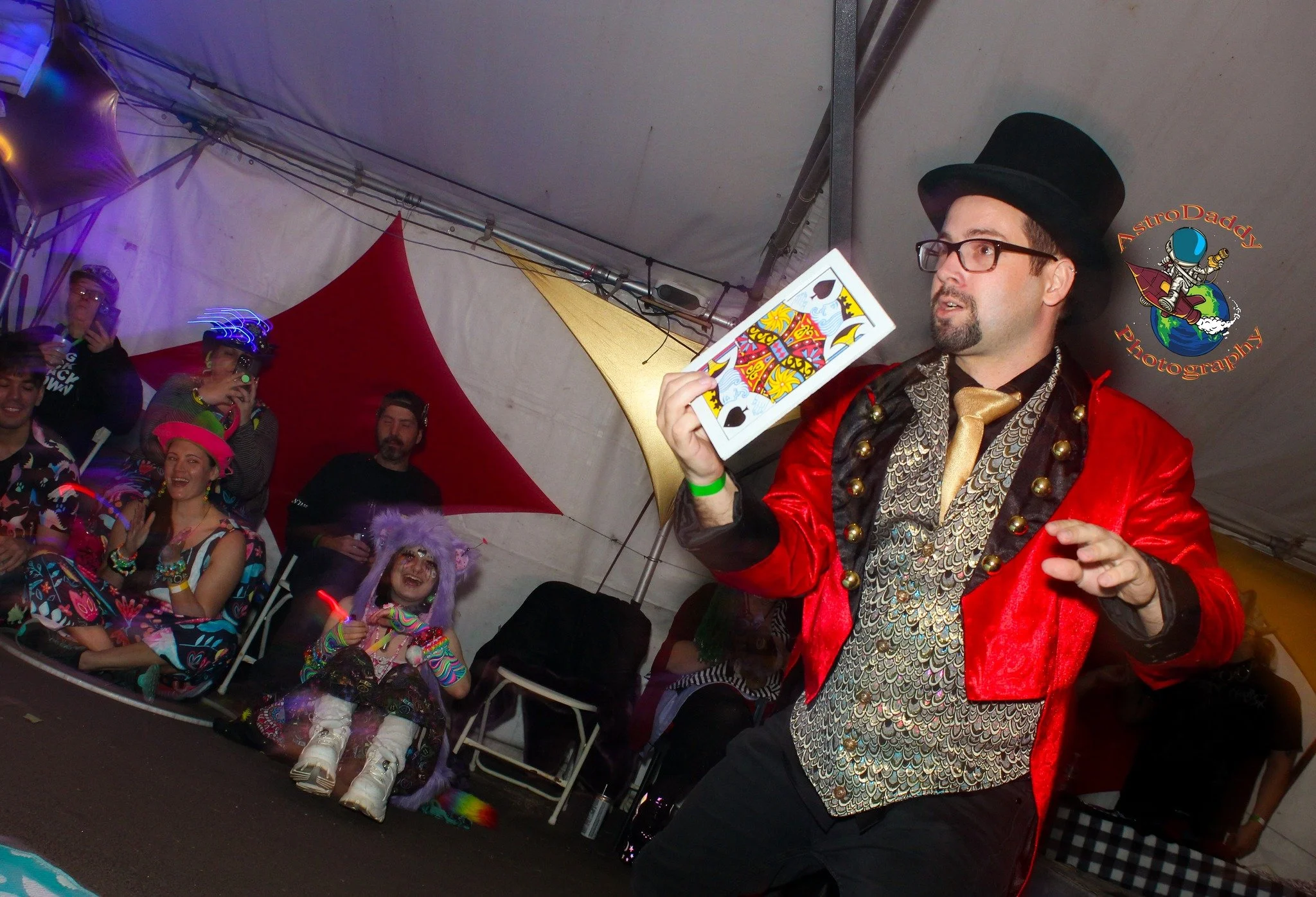 Man wearing a top hat, glasses, and a red and gold jacket performs a card magic trick in front of an audience dressed in colorful costumes.