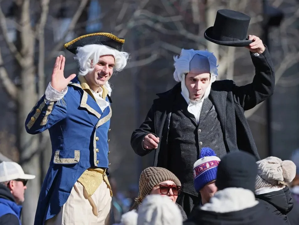 Two men dressed as historical figures, one in a blue coat and the other in black, interacting with a crowd during an outdoor event. The man in blue is waving and smiling, wearing a white wig and a tricorn hat, suggesting a colonial or revolutionary era costume. The man in black is holding a top hat, also wearing a white wig, and appears to be speaking.