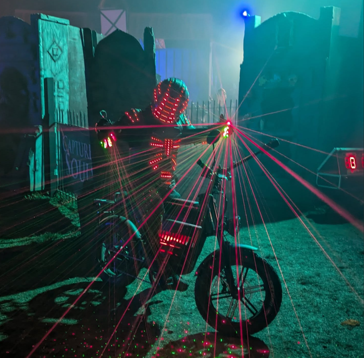 A person in a costume with LED lights, standing next to a bike with LED decorations, and laser lights.
