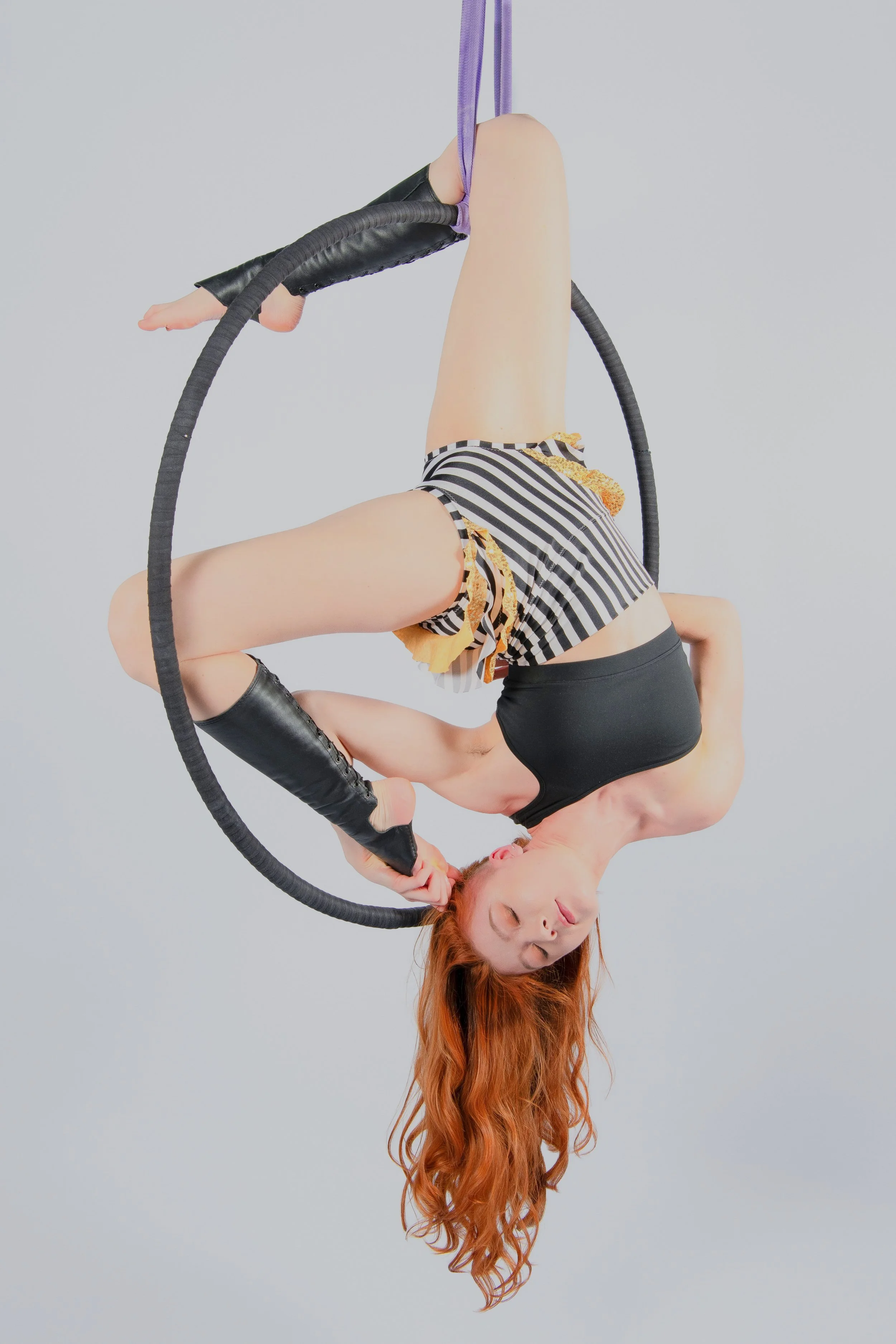 A woman with long red hair performing an aerial hoop act, hanging upside down with her eyes closed, wearing a black tank top, striped shorts, and black knee-high lace-up boots.