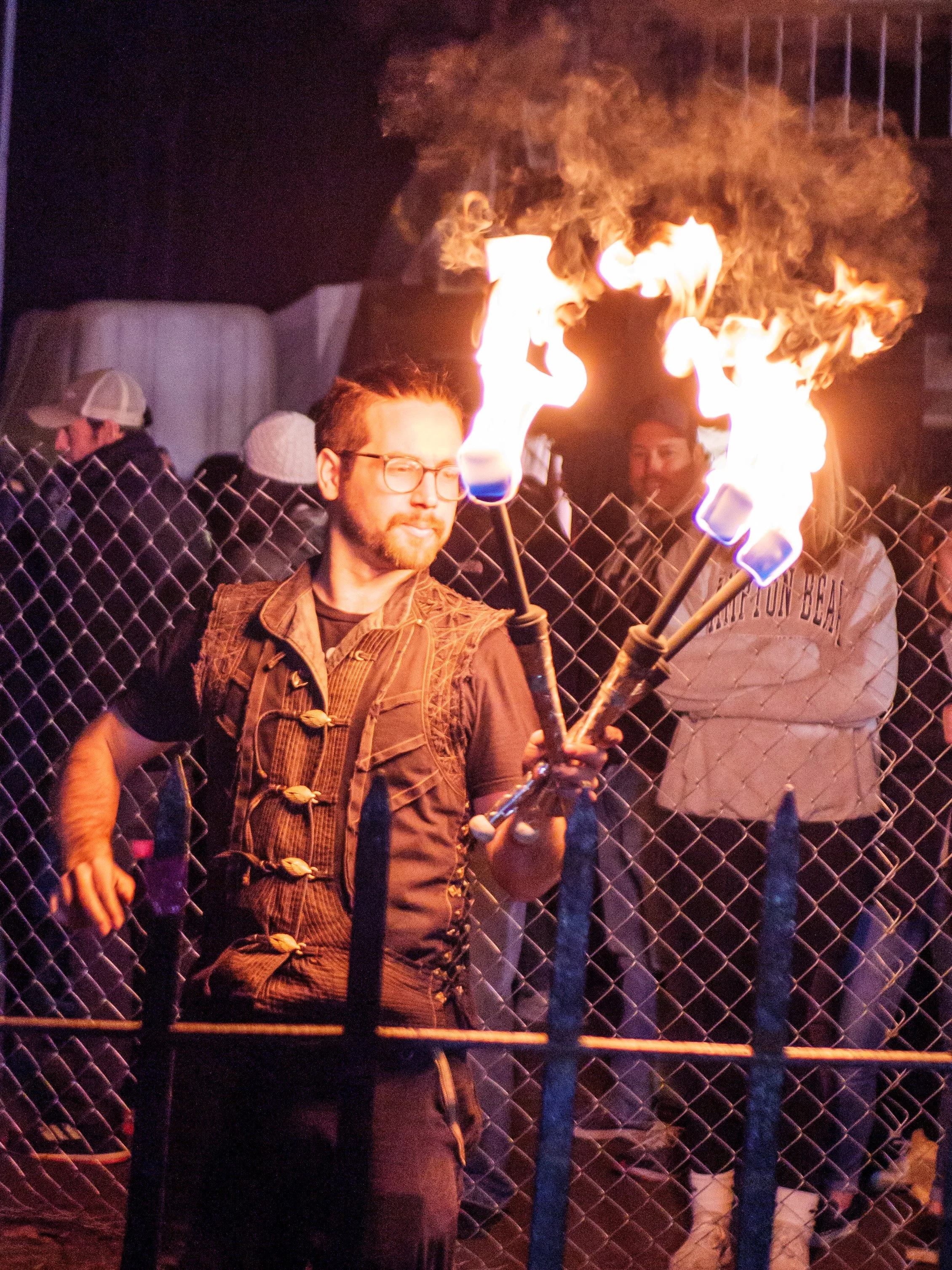 A man in glasses and a black outfit performs fire juggling at night, with a chain-link fence and spectators in the background.