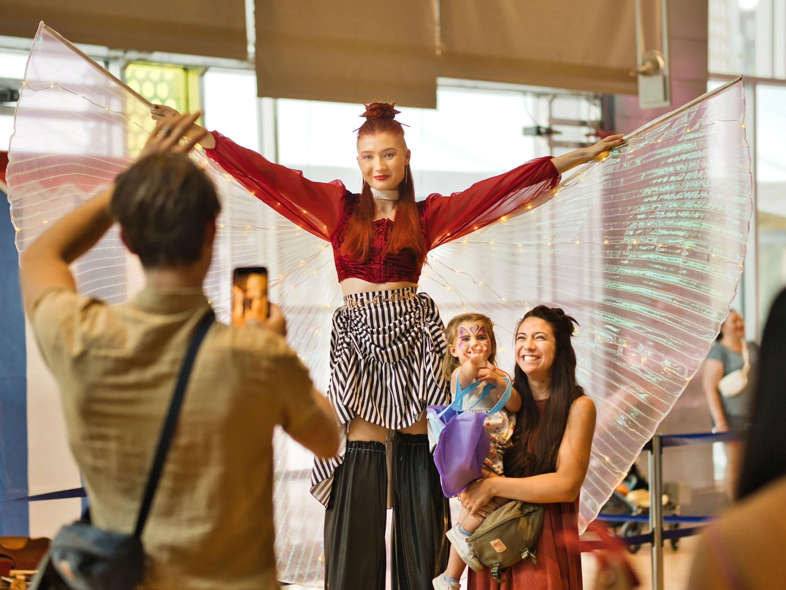 A stilt walker dressed in a red crop top and striped skirt standing with large iridescent wings, holding her arms out, while a woman and a young girl pose smiling in front of her. A person is taking a photo of them with a smartphone.