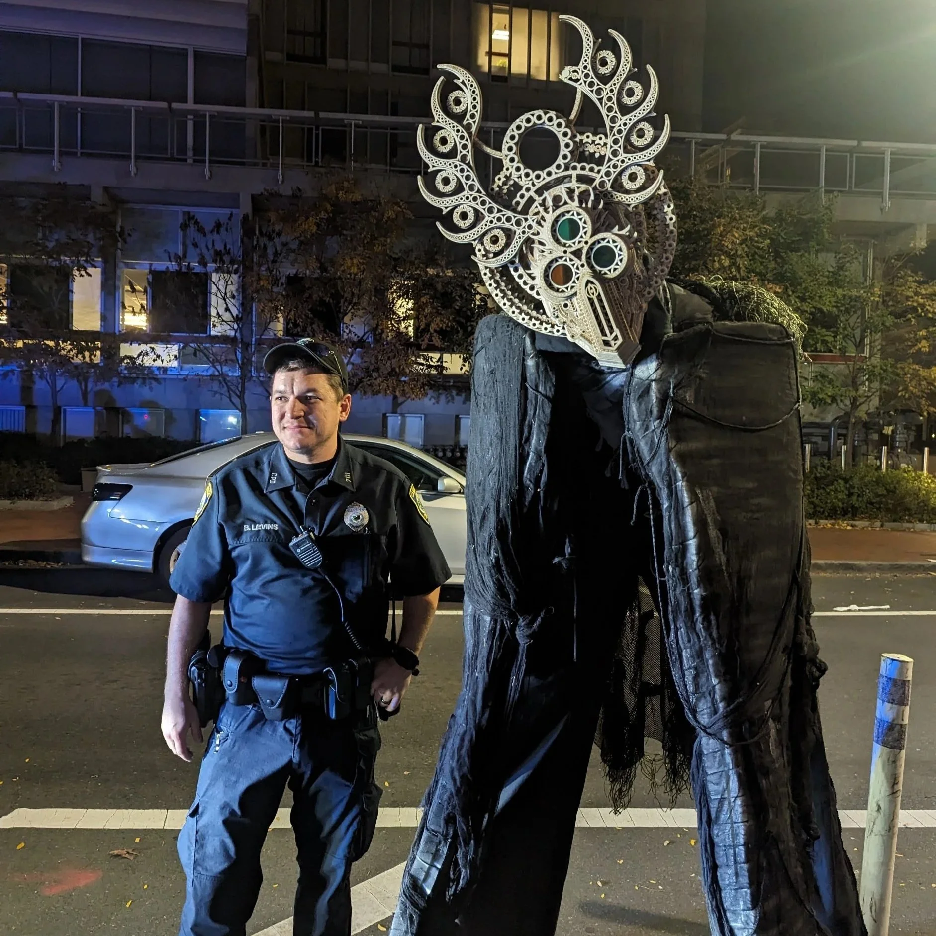 A police officer standing next to a person dressed in a dark costume with an intricate mask, on a city street at night.