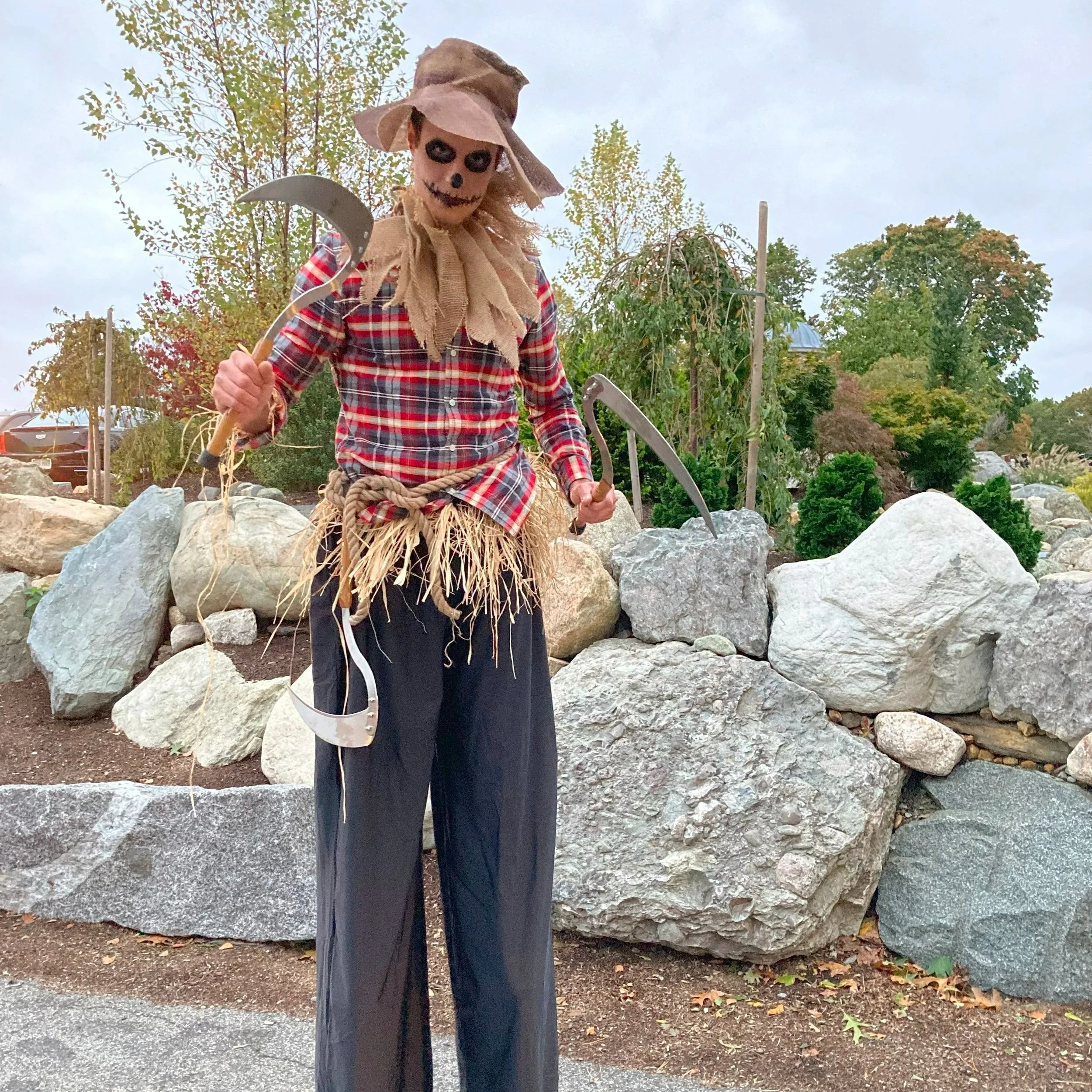 A person dressed as a scarecrow with a skeleton face, wearing a wide-brimmed hat, plaid shirt, straw skirt, and black pants, holding two sickles in an outdoor garden area with rocks and trees.