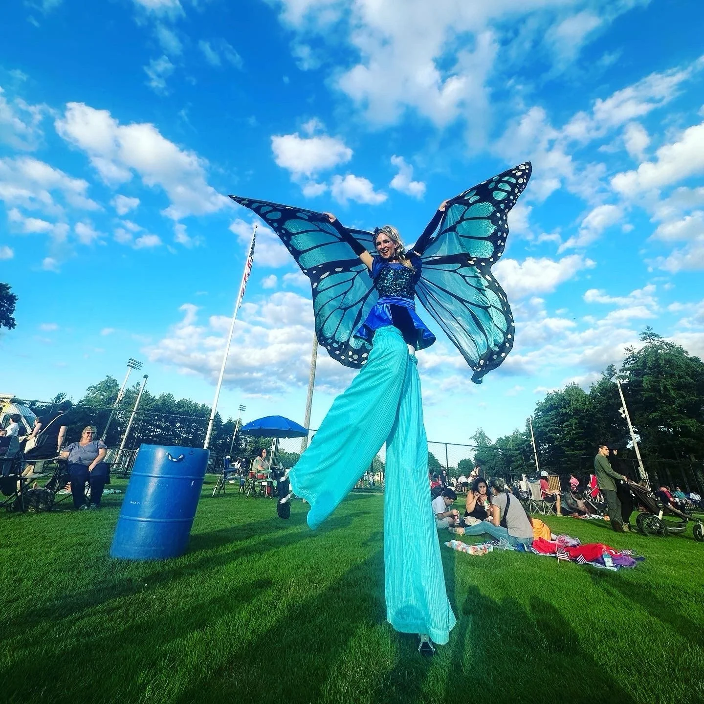 A performer on stilts dressed as a butterfly with large blue wings and long flowing blue skirt, performing for a crowd in an outdoor park on a sunny day with a partly cloudy sky.