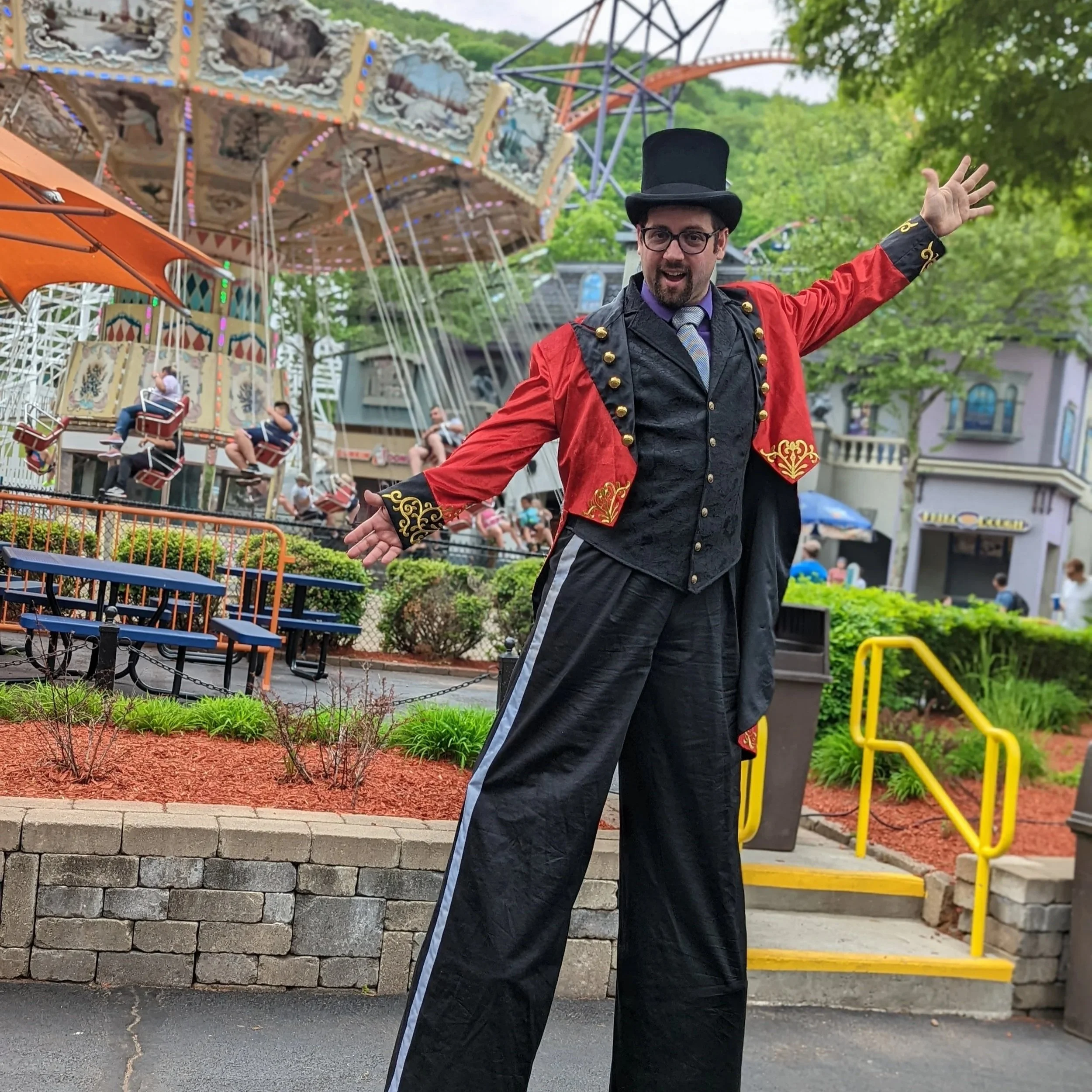 A man dressed as a circus or carnival ringmaster with a black top hat, red coat with gold details, and oversized pants, standing with arms outstretched in front of a carousel with swings and riders, and an amusement park with people in the background.