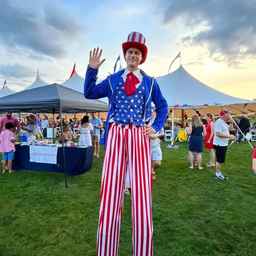 A man dressed as Uncle Sam on stilts waving at a festive outdoor event with tents, people, and decorations in the background.