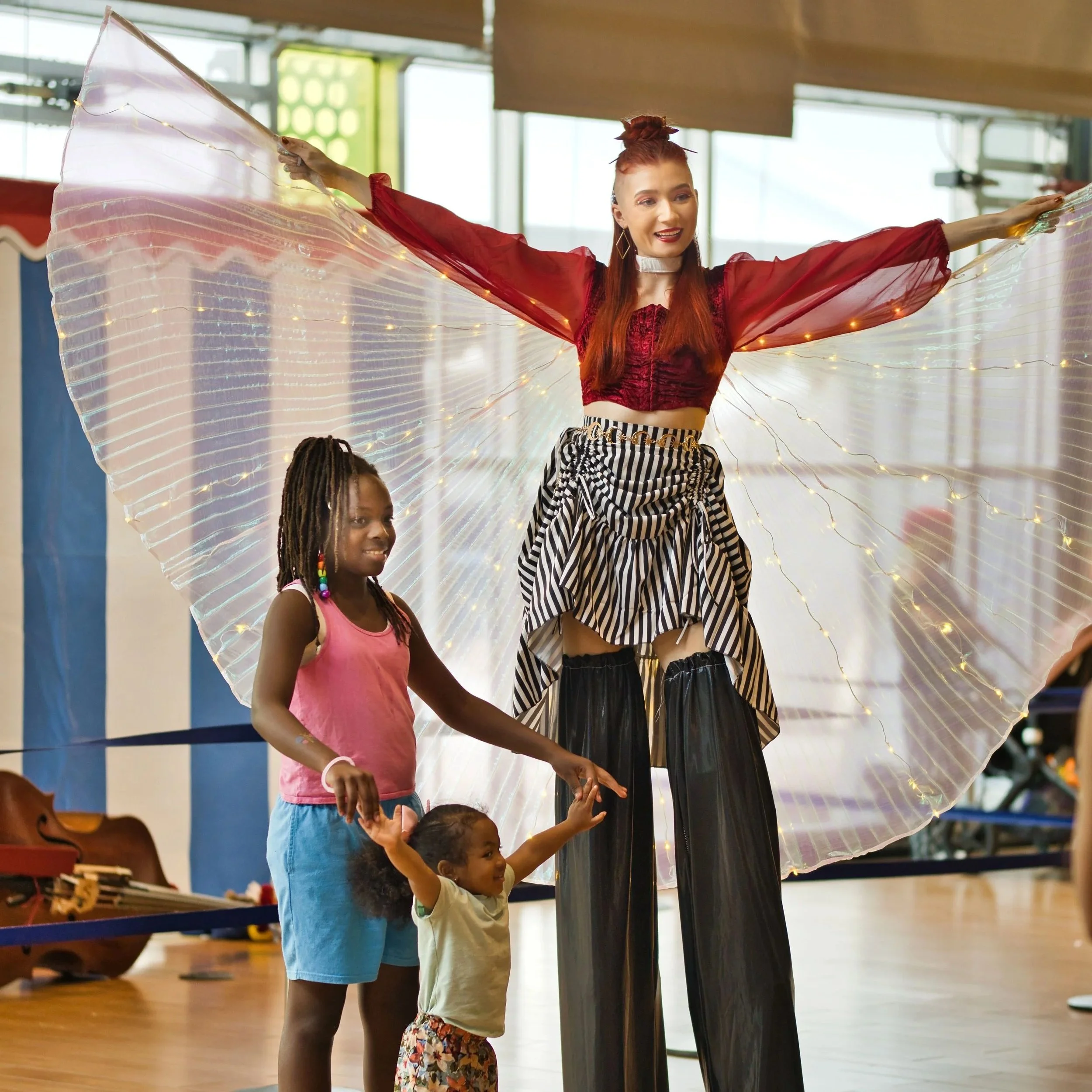 A woman on stilts dressed in colorful, theatrical clothing, with strappy wings illuminated with small lights, in a spacious indoor setting. Two young girls are next to her, one smiling and reaching up, the other looking up with excitement.