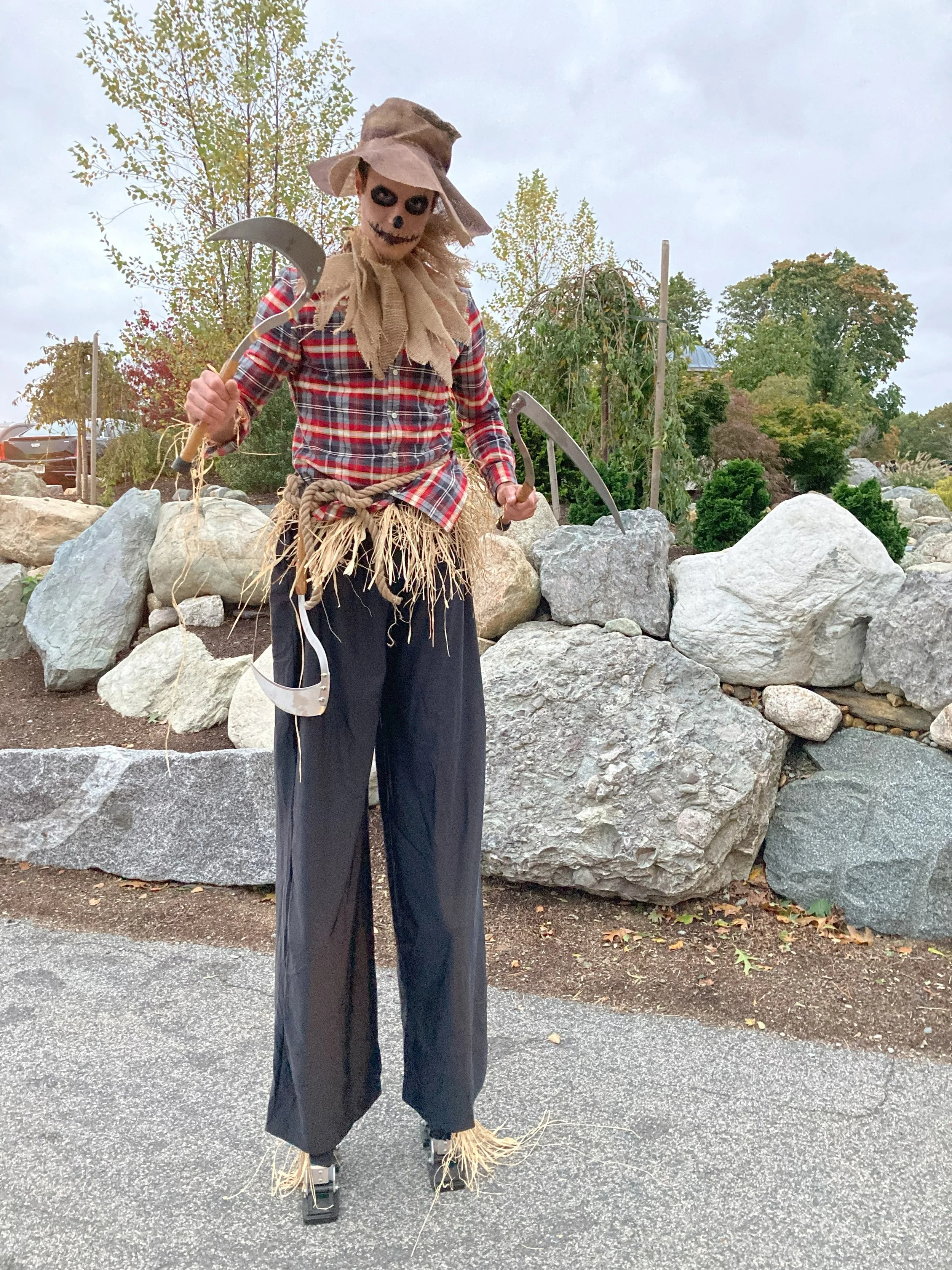 A scarecrow with a skeletal face, black eye makeup, wearing a plaid shirt, straw skirt, and a wide-brimmed hat, holding sickles, and standing outside near rocks and trees.