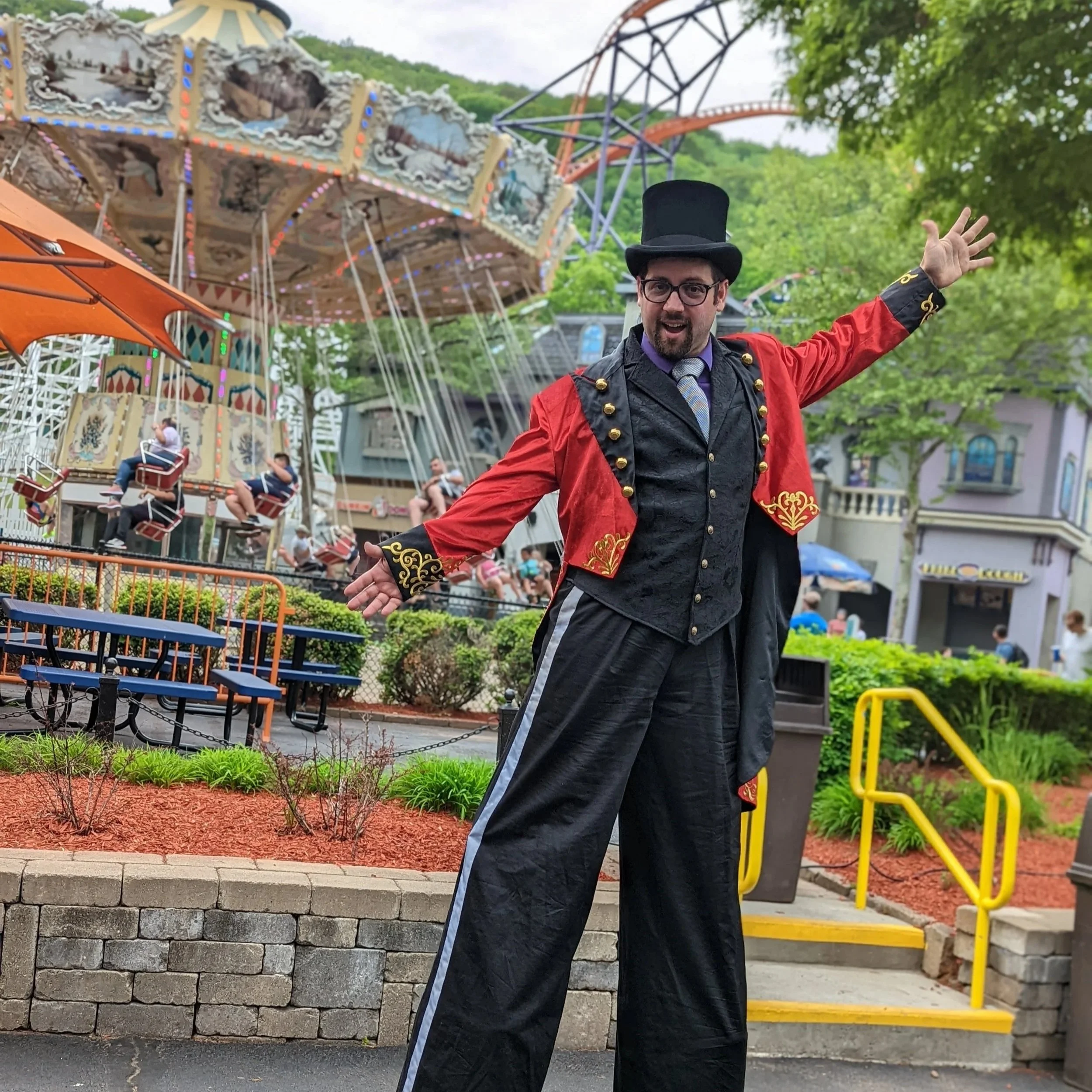 A man dressed as a circus ringmaster with a black top hat, red jacket with gold embroidery, black pants, and a vest, stands with arms open in front of a carousel ride at an amusement park.