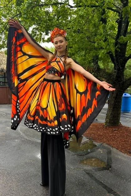 A woman dressed in a butterfly costume with orange, yellow, and black wings, and a matching headpiece, standing outdoors on a paved area near trees.