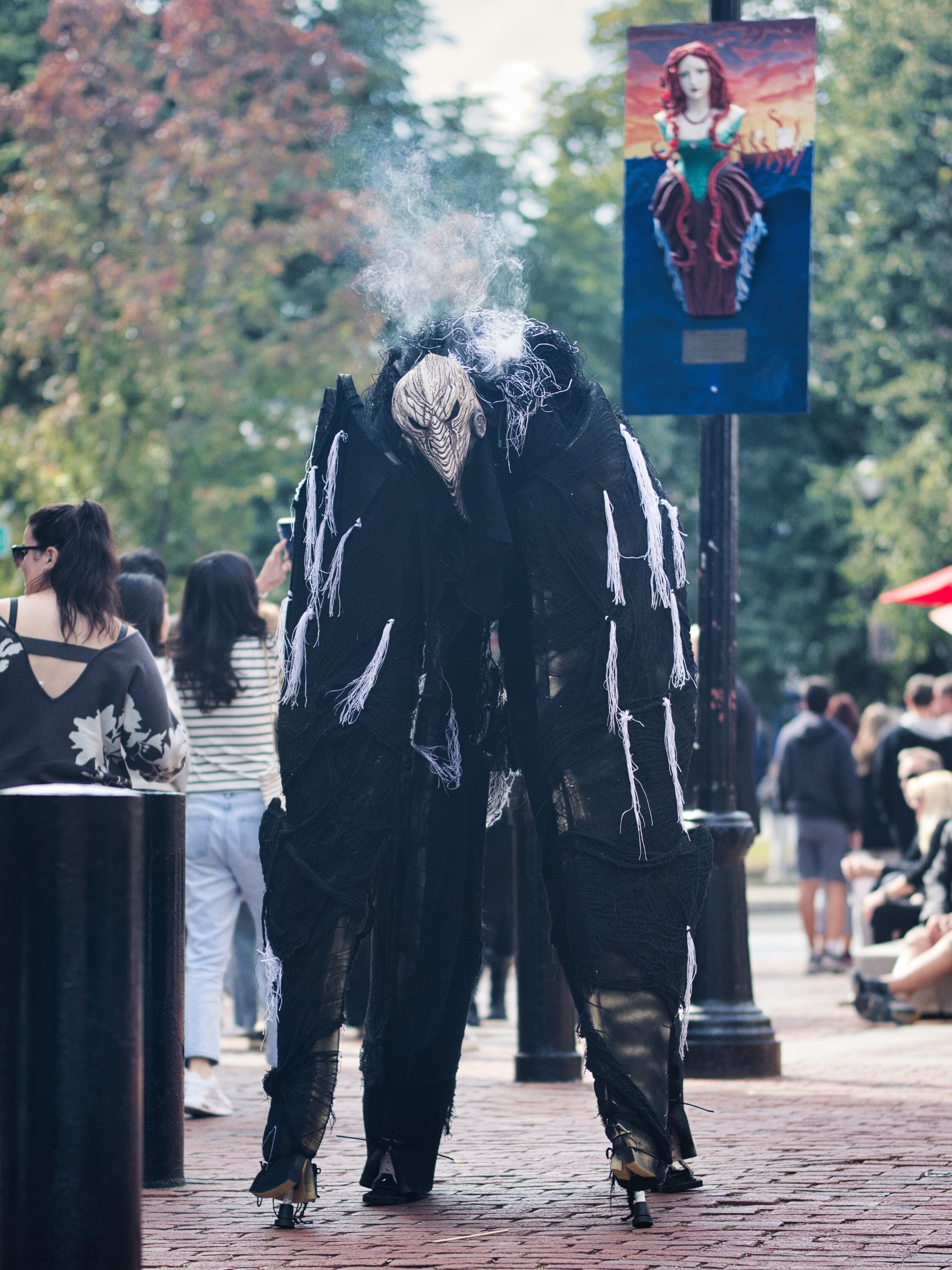 Person dressed in a black costume with a wooden mask and cigarette smoke, surrounded by people at an outdoor event with trees and a colorful artwork in the background.