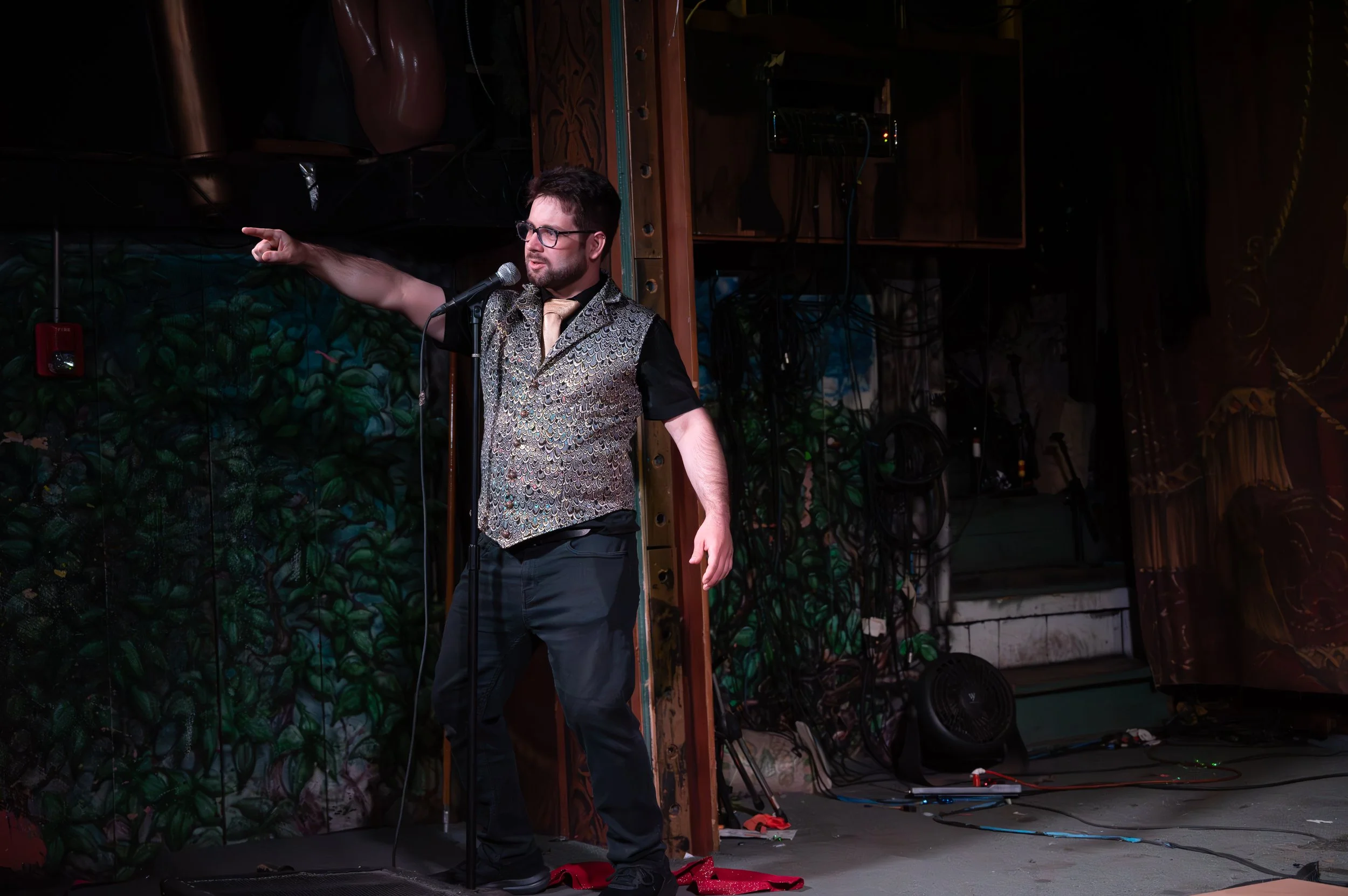 A man with dark hair, glasses, and facial hair stands on stage. He is wearing a patterned vest over a black shirt, with a tan tie. He appears to be performing, with his left arm extended outward, pointing to his left, in front of a colorful backdrop 