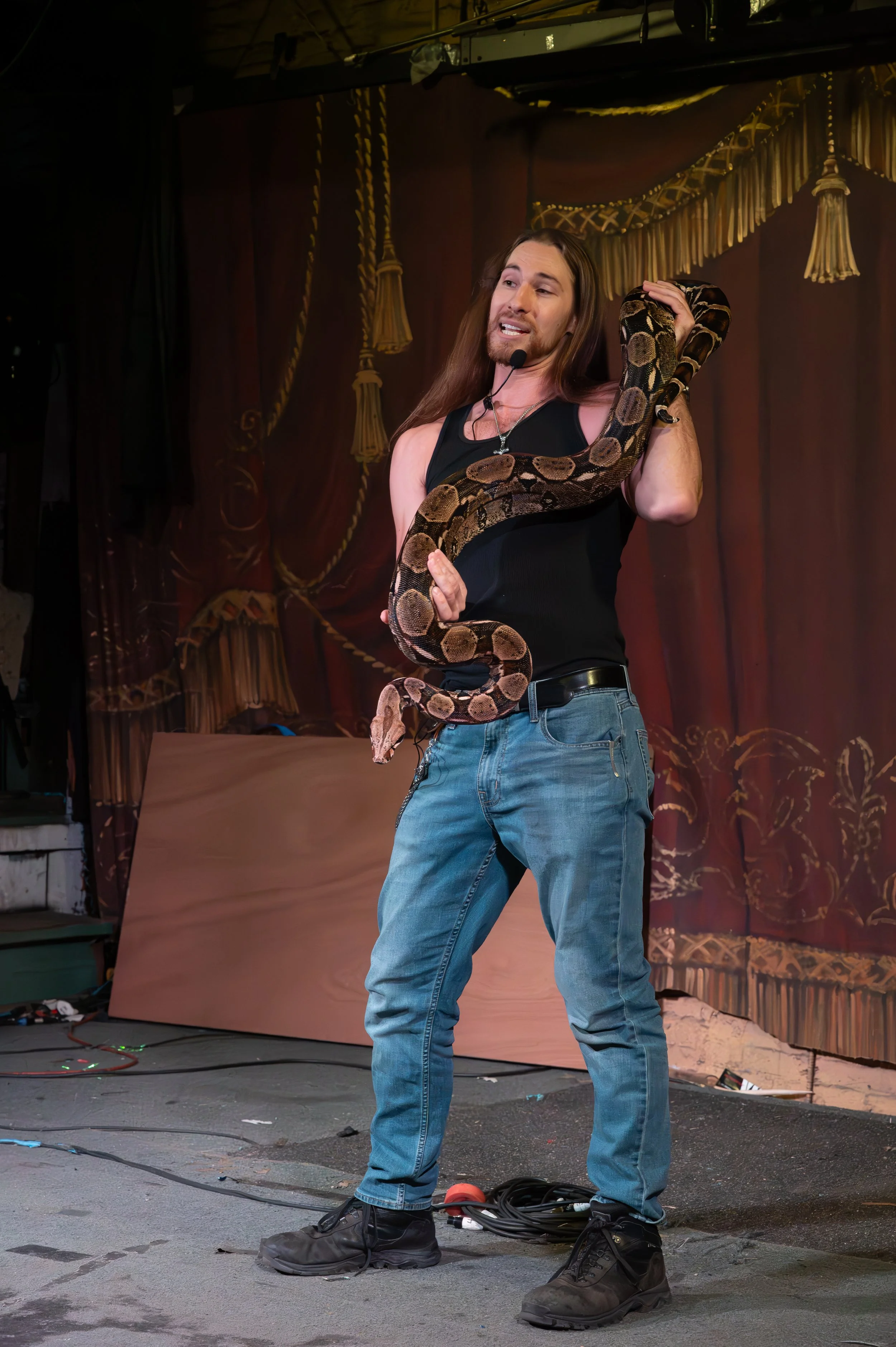 A man with long hair, wearing a black tank top, blue jeans, and black shoes, holding a snake on stage with a red curtain backdrop.