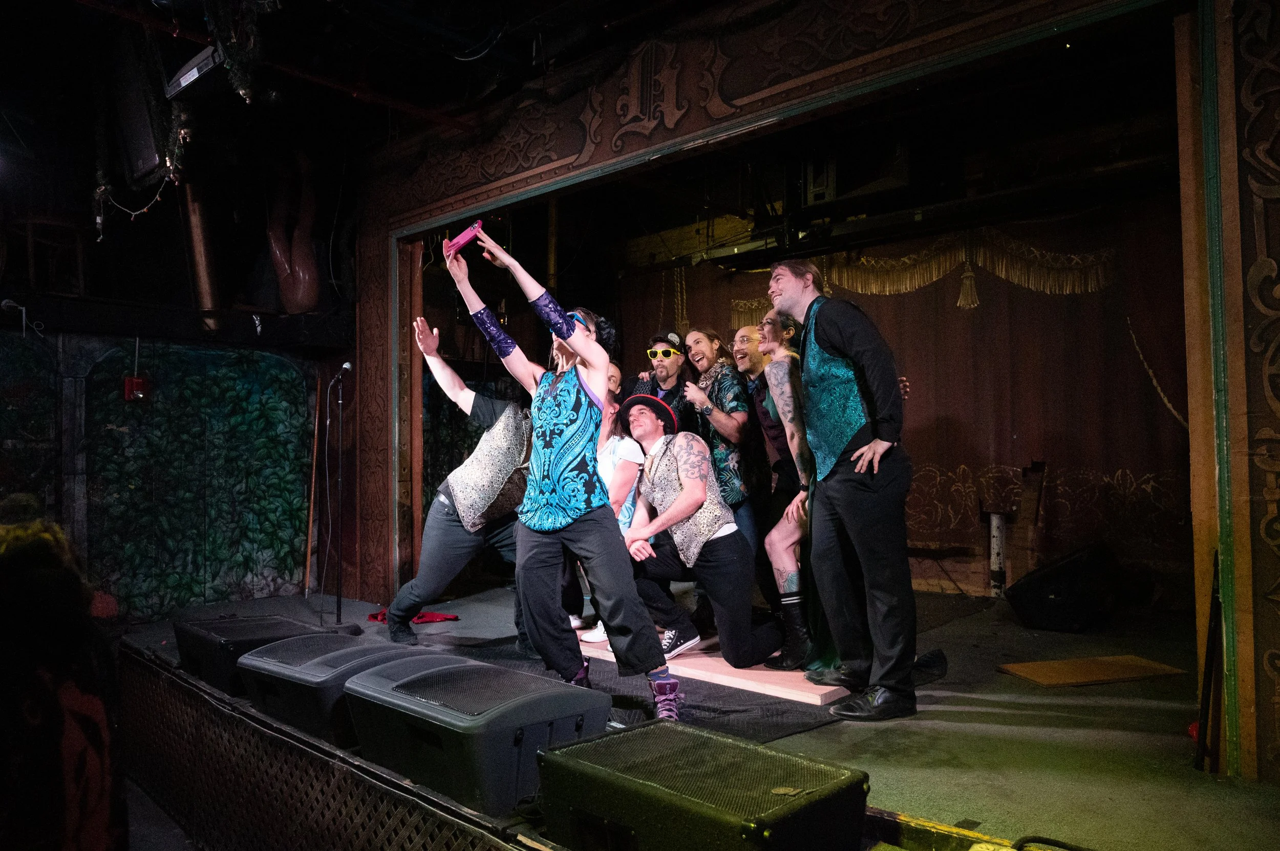 A group of people on stage taking a selfie and posing for a photo, dressed in colorful clothing and costumes, with stage equipment and microphones visible.