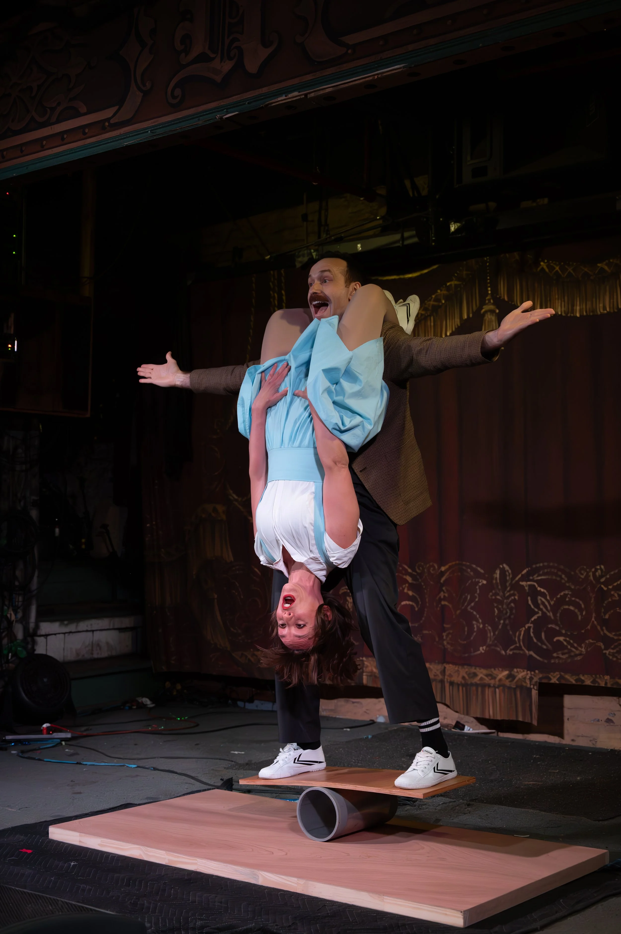 A man in a brown blazer and black pants balancing on a wooden board placed on a cylindrical object, while a woman in a light blue dress is upside down with her head near the man's waist, holding onto him with her hands, in a theatrical or comedy perf